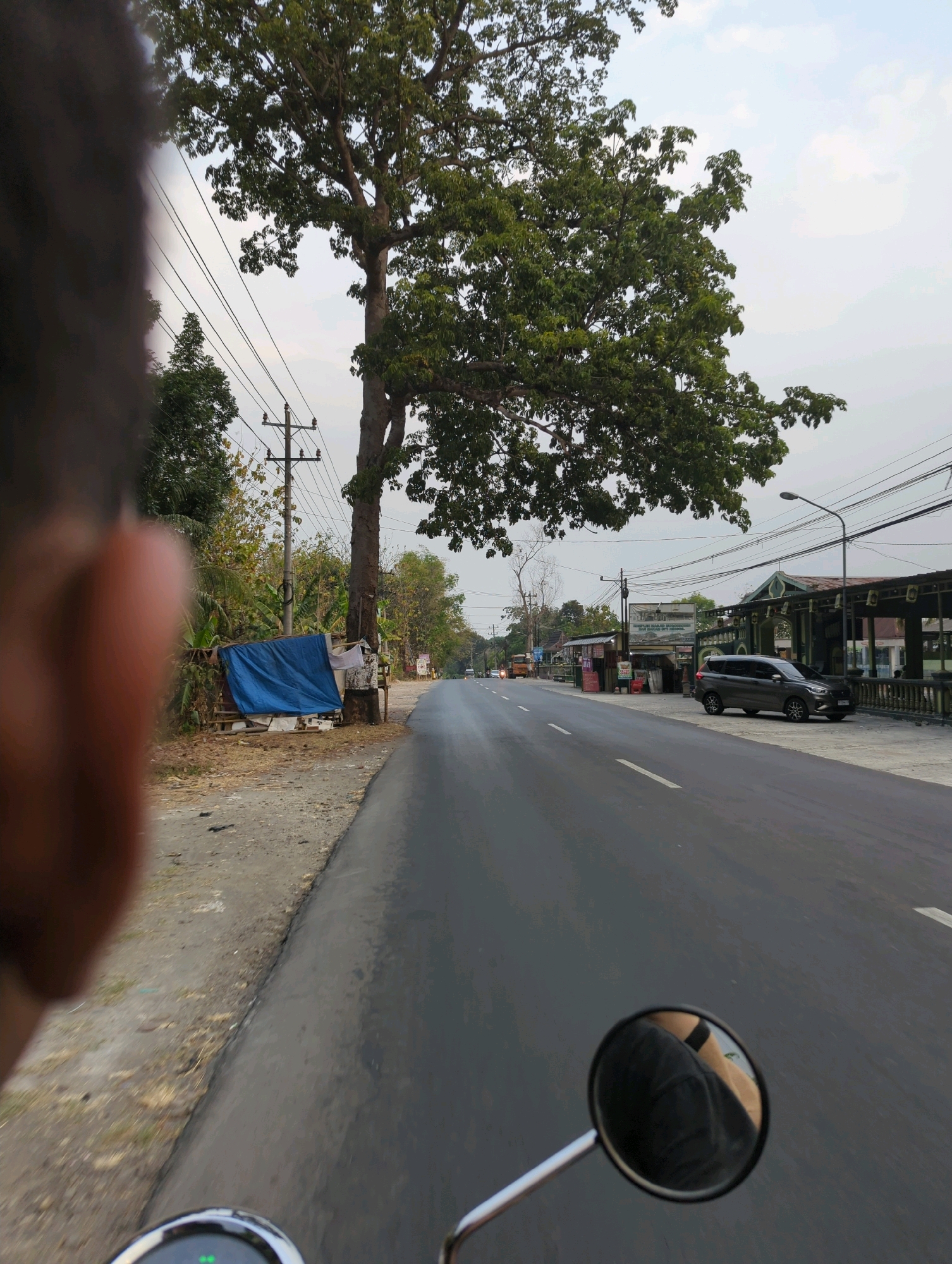 The image depicts a view from the perspective of someone riding a motorcycle through a rural road in Kacangan, Indonesia. On the left side of the road, there is a large tree with lush green foliage, a utility pole with multiple wires, and a small roadside stall covered with a blue tarp. The road itself is asphalt and appears wet, suggesting recent rain or morning dew. In the background, there are buildings and more trees, hinting at a small town or village. On the right side of the road, there is a parking area with a dark-colored SUV. A street lamp is visible above the buildings. The sky is overcast with a pale hue. In the foreground, a motorcycle's rearview mirror reflects the rider's leg and clothing. The overall impression is a snapshot of daily life on a road in Indonesia, with a blend of natural and man-made elements.