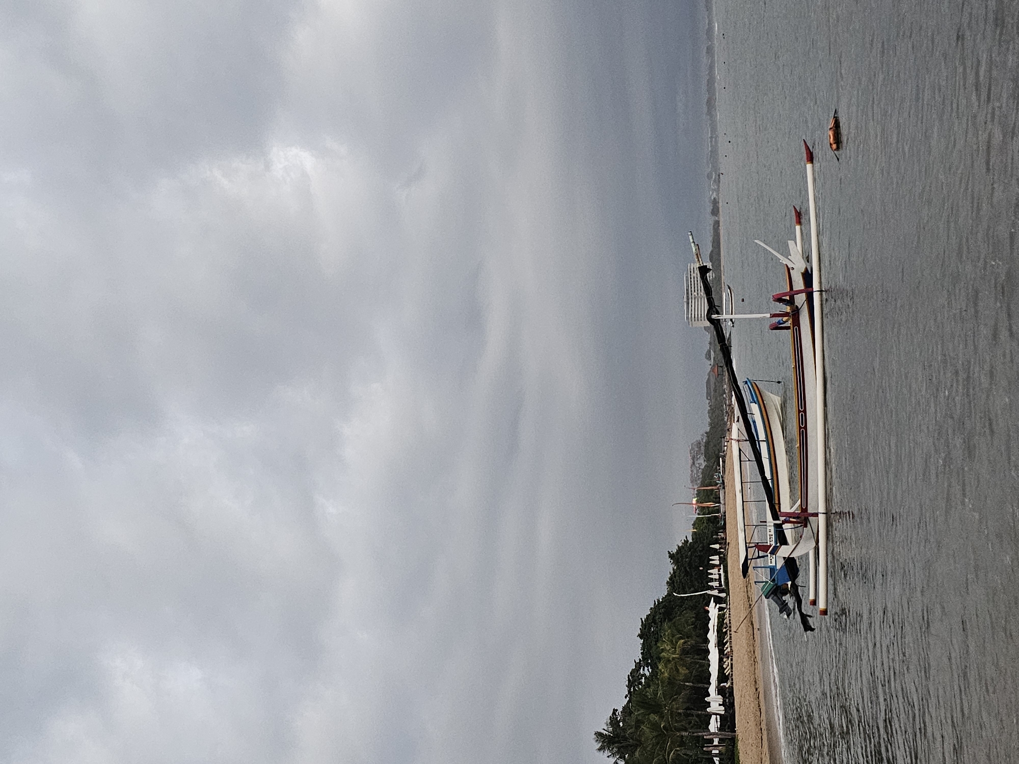 The image depicts a beach scene in Denpasar, Indonesia under an overcast sky. The predominant feature is a traditional outrigger boat, known as a Jukung, resting on the shallow water's edge. The boat is painted in white, red, and blue, with supporting bamboo or wooden structures extending outwards. A small outboard motor is attached to the back of the boat. A small orange buoy is floating in the water nearby.
The shoreline is lined with palm trees and other vegetation, indicating a tropical environment. Several other similar boats are pulled up onto the sandy beach in the distance. The water is relatively calm.