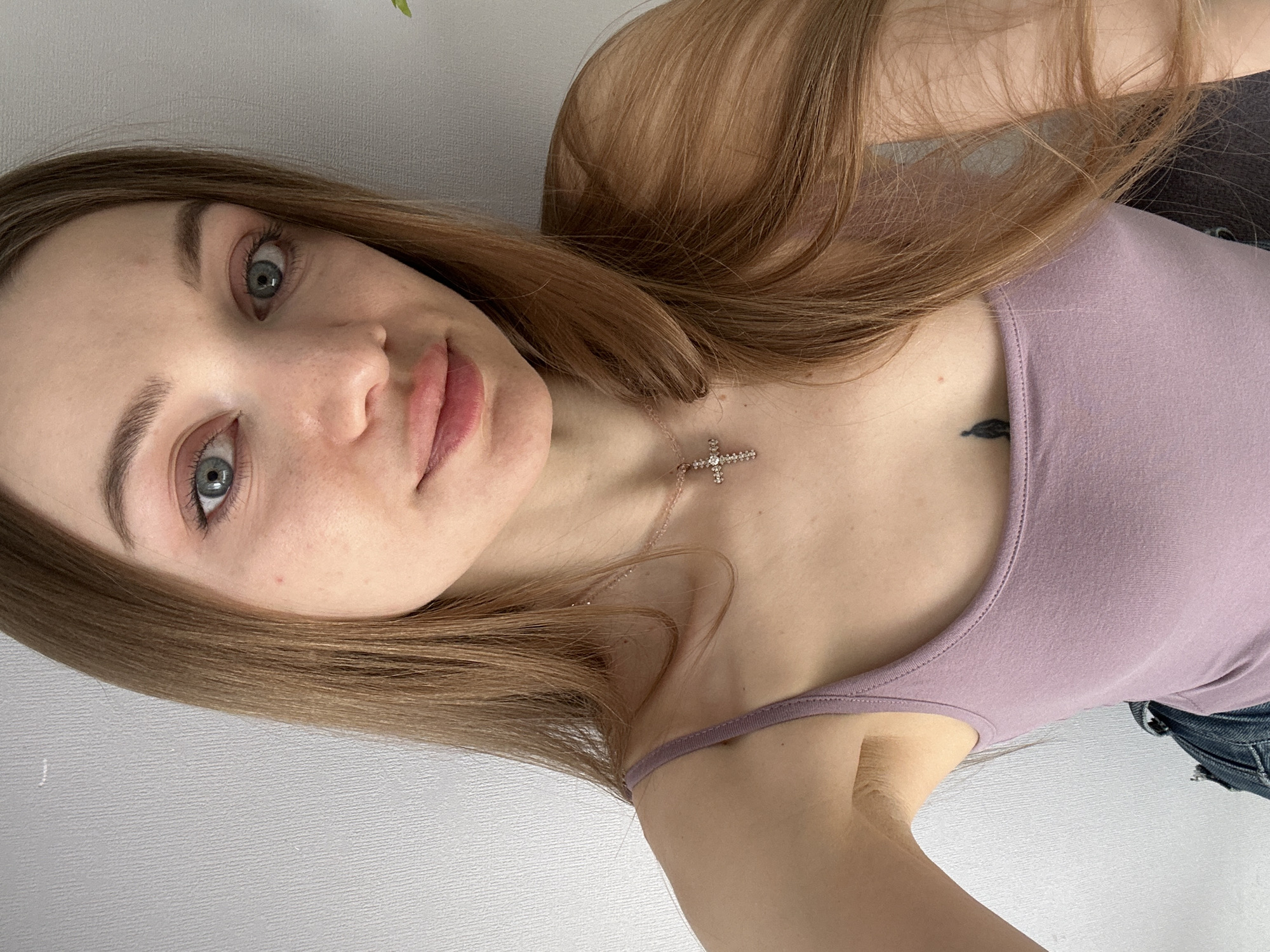A young woman is posing indoors. She has long, light brown hair and blue eyes. She is wearing a light purple tank top and a cross necklace. There is a small black tattoo on her chest. The background is a plain white wall. There is no indication of the city of Kazan, Russia.