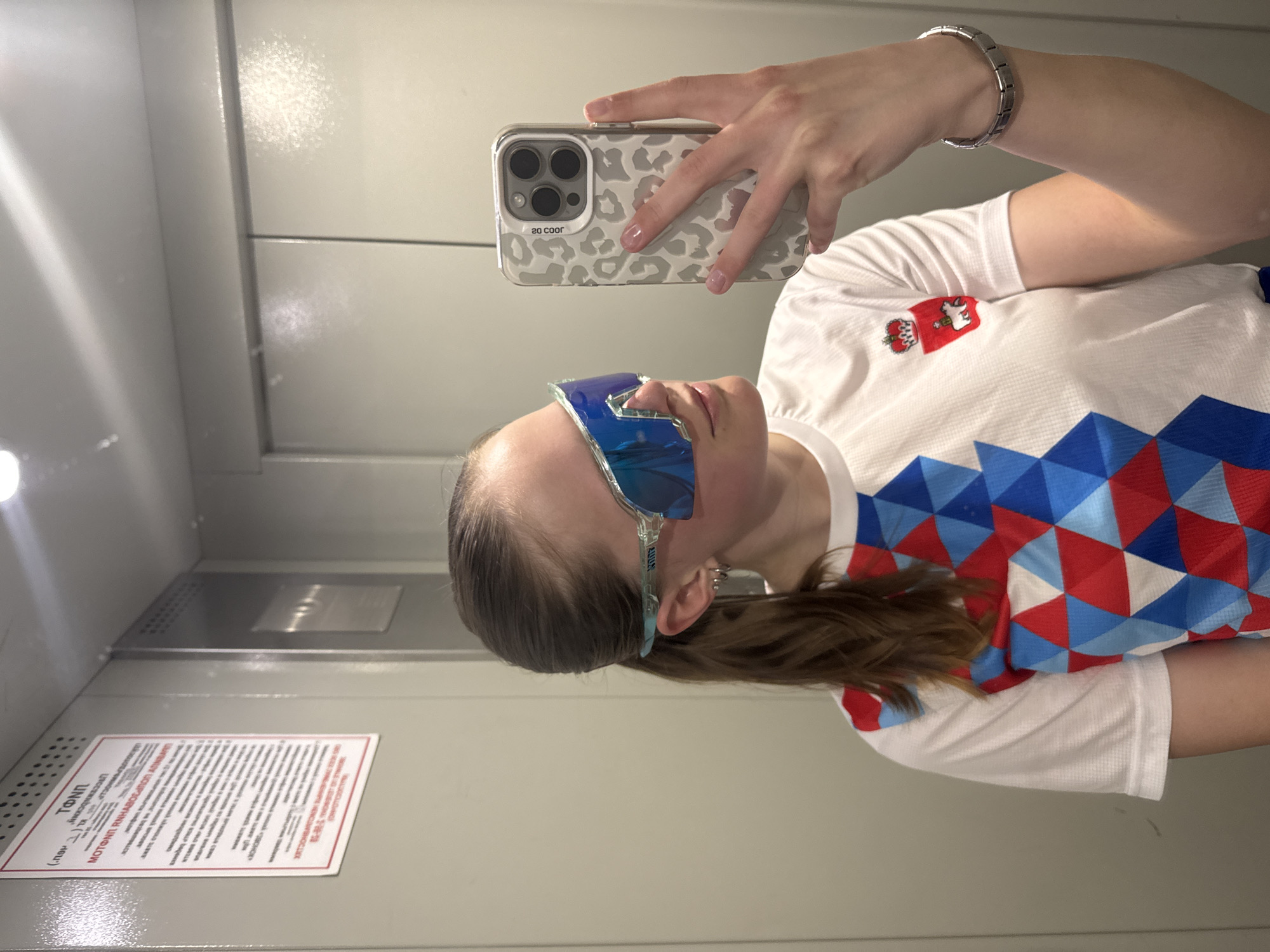 A person takes a selfie in an elevator. The individual is wearing reflective blue sunglasses and a white T-shirt with red, white, and blue geometric patterns and a coat of arms near the shoulder. The person is holding a phone with a spotted case. A silver bracelet is visible on the wrist. A notice is posted on the elevator wall, written in a non-English language. The location is an elevator in Chaikovsky, Russia.