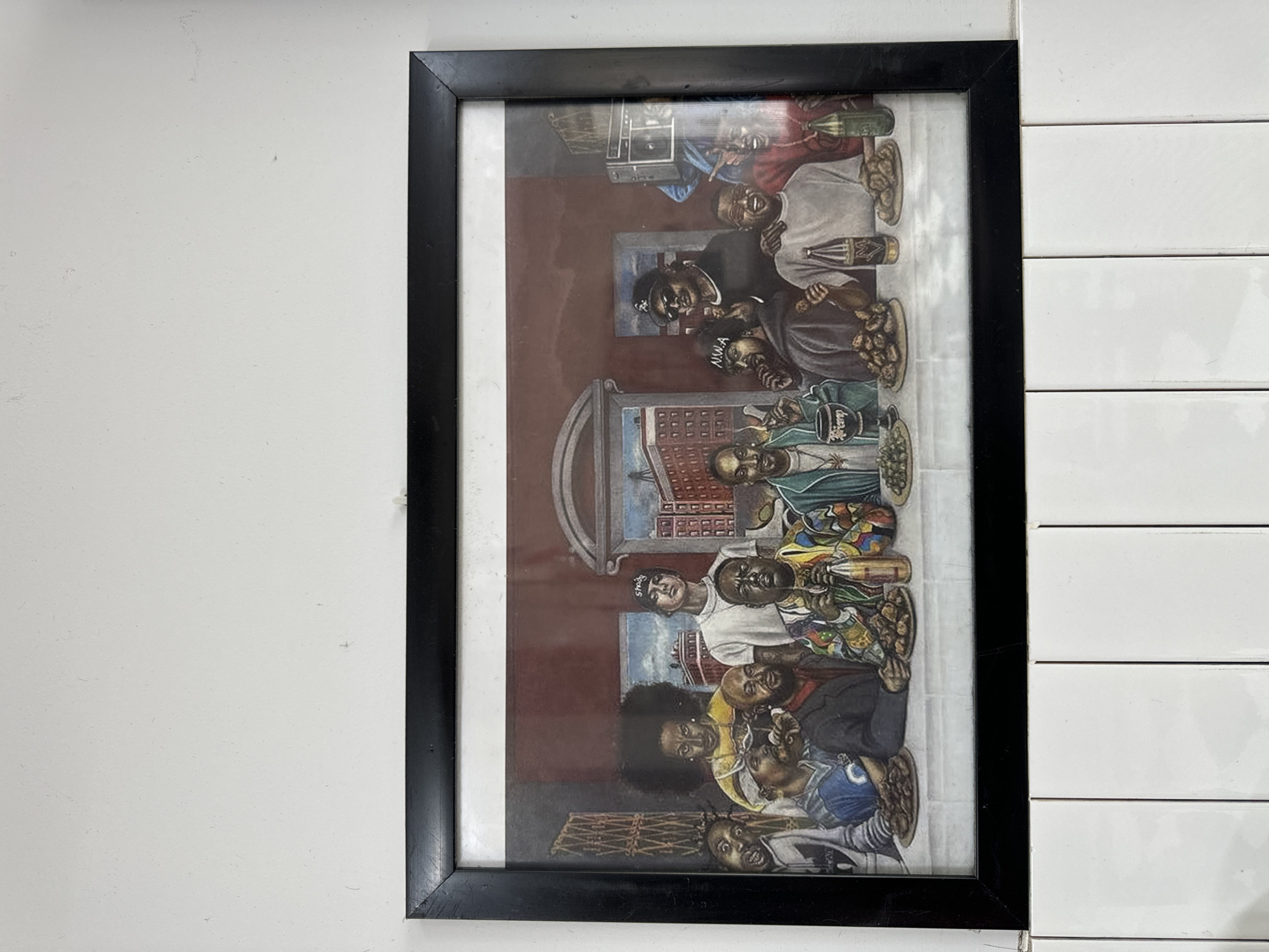 This is a photograph of a framed painting hanging on a white wall above a white tiled surface. The painting depicts a scene reminiscent of Leonardo da Vinci's "The Last Supper," but with a gathering of prominent figures from hip-hop culture. The figures are seated at a long table laden with food and drinks, and they are rendered in a realistic artistic style. The painting is a horizontal rectangle with a black frame. The white wall appears to be textured, and there are some faint marks or imperfections on its surface. The white tiled surface below is composed of rectangular tiles arranged vertically, with visible grout lines. The lighting suggests the scene is indoors, and there are no other contextual elements in the photograph that would indicate a specific location within Phan Thiết, Vietnam.