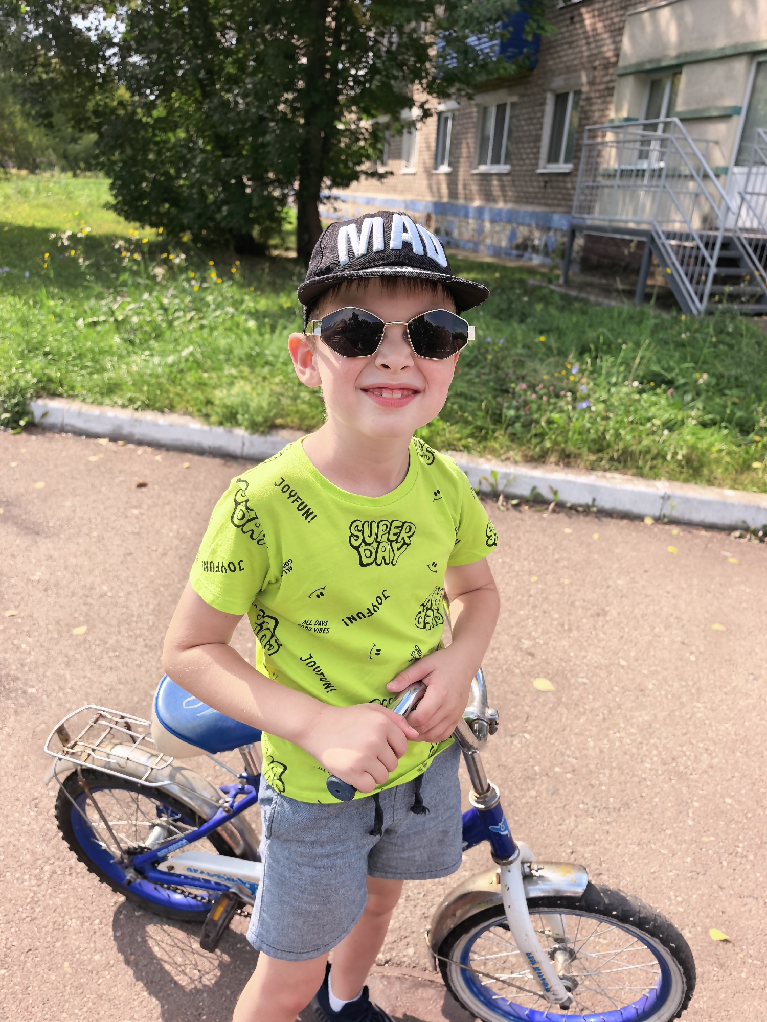In a sunlit outdoor setting, a young boy stands next to a blue and white bicycle. He is wearing a black baseball cap with white lettering that reads "MAN", mirrored sunglasses, a bright lime green t-shirt with various graphic prints and lettering, and grey shorts. The boy is smiling and holding the handlebars of the bicycle. The bicycle has a blue seat, a silver rack over the rear wheel, and a blue and white frame. The ground is paved with asphalt, and there are some yellow leaves scattered on it. In the background, there is a grassy area with scattered wildflowers and a multi-story building with windows and an external staircase. The environment suggests a residential area in the city of Sterlitamak, Russia, likely a courtyard or a street.