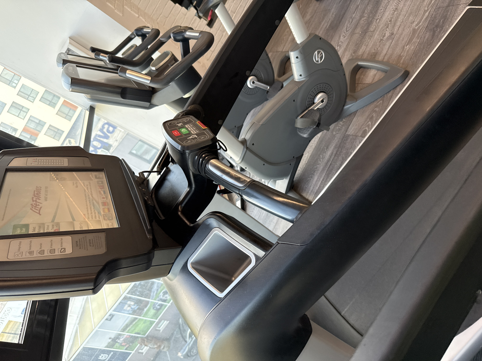 The image shows a close-up view of a Life Fitness treadmill in what appears to be a gym setting. The treadmill screen displays workout options, and part of a building can be seen through the window in the background. Adjacent to the treadmill are stationary bikes. The location could be in the city of Sofia, Bulgaria, as indicated by the OCR text in the image.