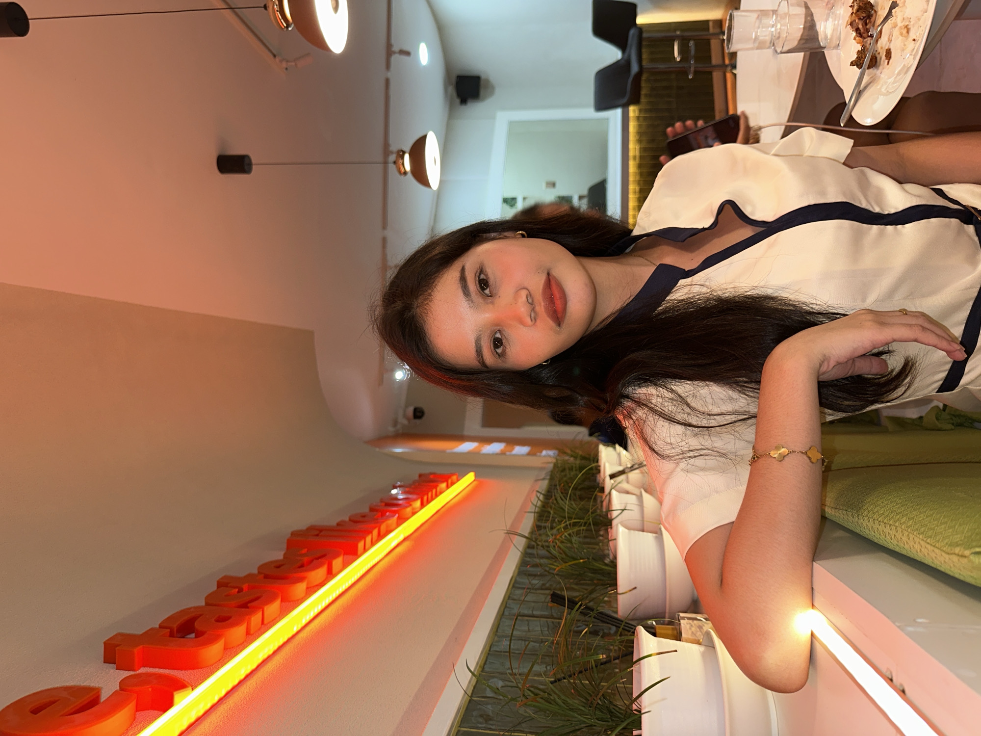 The image shows a young woman sitting at a table in what appears to be a restaurant or cafe. She has long dark hair and is wearing a light-colored blouse with dark trim. Her left arm is resting on a surface, and she is wearing a bracelet. In the background, there are plants in white pots, a neon sign with red lettering that appears to say "taste like", and a bar area with stools. The overall lighting suggests an indoor evening setting. The presence of a neon sign that reads "taste like" in conjunction with the restaurant setting strongly suggests that the location is likely "Taste Like" restaurant, a known establishment in Davao City, Philippines. The woman is looking towards the camera. There are plates with food and glasses on the table in front of her.