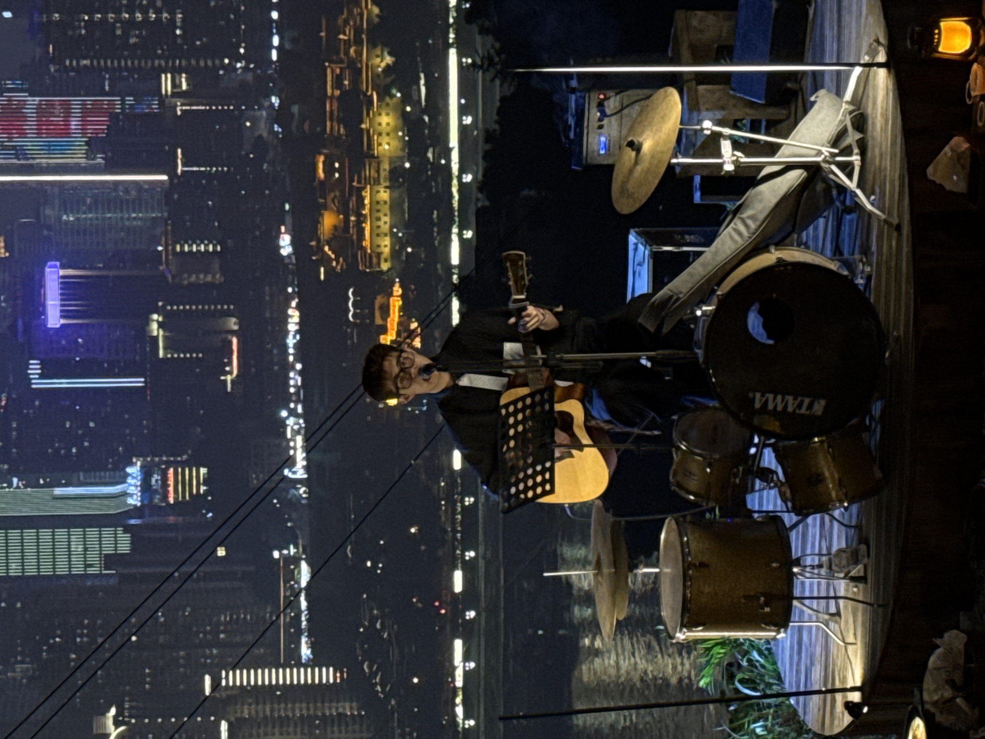 A musician plays guitar and sings on an outdoor stage. He wears glasses, a white dress shirt, and a black jacket. A music stand is in front of him, and a drum set is to his right. The background shows the night skyline of Nan'an District, China, with lit-up buildings. Overhead lines are also visible in the foreground.