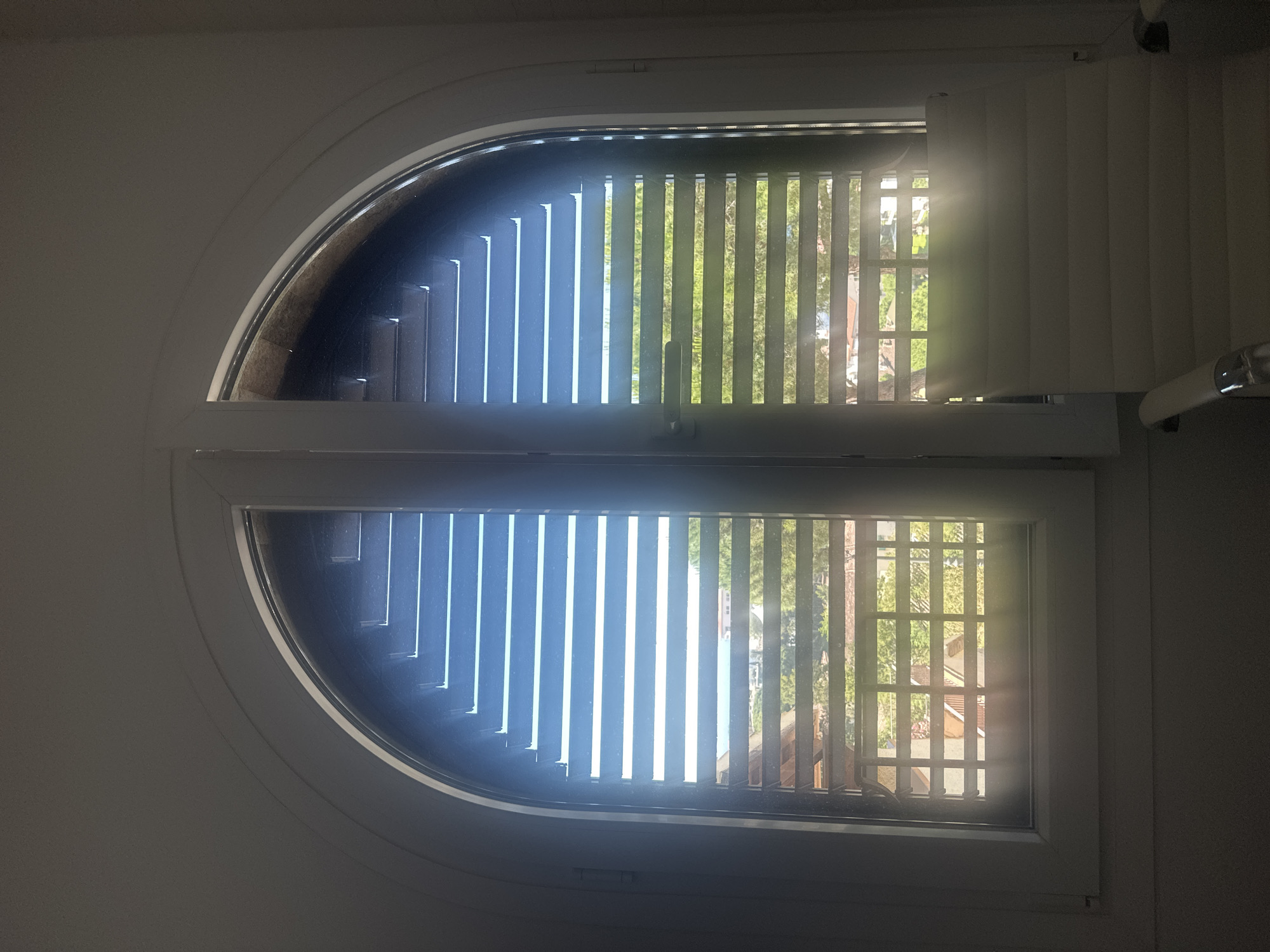 A window with vertical blinds is shown in an interior space. The window is uniquely shaped, with a rounded top half and a rectangular bottom half. The blinds are mostly closed on the left side, with some light filtering through. On the right side, the blinds are more open, revealing a view of trees and buildings, indicative of an urban or suburban setting. The window frame is white. The image is rotated. There is no information on this image to suggest its location is in Sant Feliu de Guíxols, Spain.