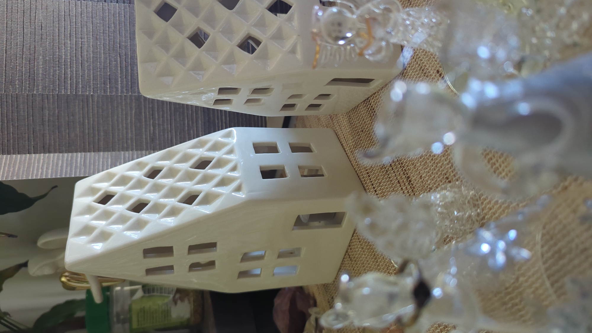 In the city of Kløfta, Norway, the image captures a close-up view of two white ceramic decorative houses with diamond-shaped cutouts on their roofs. The house on the left is more in focus, showing square window-like cutouts on its sides. To the right, another similar ceramic house is partially visible. In the foreground, several out-of-focus, translucent, angel-shaped figurines are arranged, creating a soft, blurred effect with shimmering highlights. The objects are placed on a textured, light brown surface, possibly a placemat or burlap. In the background, a tall, vertically striped grey object and a hint of greenery suggest an indoor setting, possibly a shelf or cabinet. The overall lighting is soft, with some light reflecting off the glossy surfaces of the ceramic houses and the figurines.