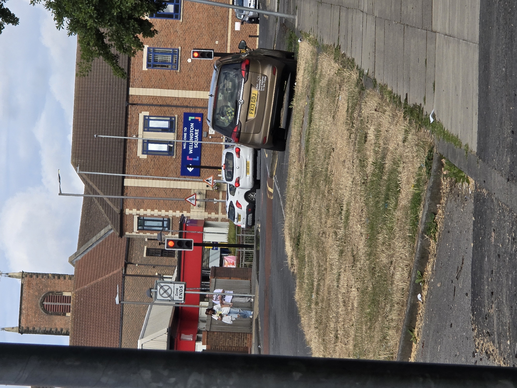 The image shows a street scene in Stockton-on-Tees, United Kingdom. In the foreground, a dry, grassy verge slopes down towards a road. A paved sidewalk runs alongside the verge. In the middle ground, a gold-colored car with the license plate "FX13 GDJ" is parked on the side of the road. A white car with license plate "HK15 HXX" is behind it. A red building with a sign that reads "WELCOME TO WELLINGTON SQUARE" is visible in the background. A traffic light, which is red, is also visible. Two individuals are walking on the sidewalk in the background, appearing to be holding phones. A brick church tower with a single arched window is to the left of the red building. The sky is partly cloudy.