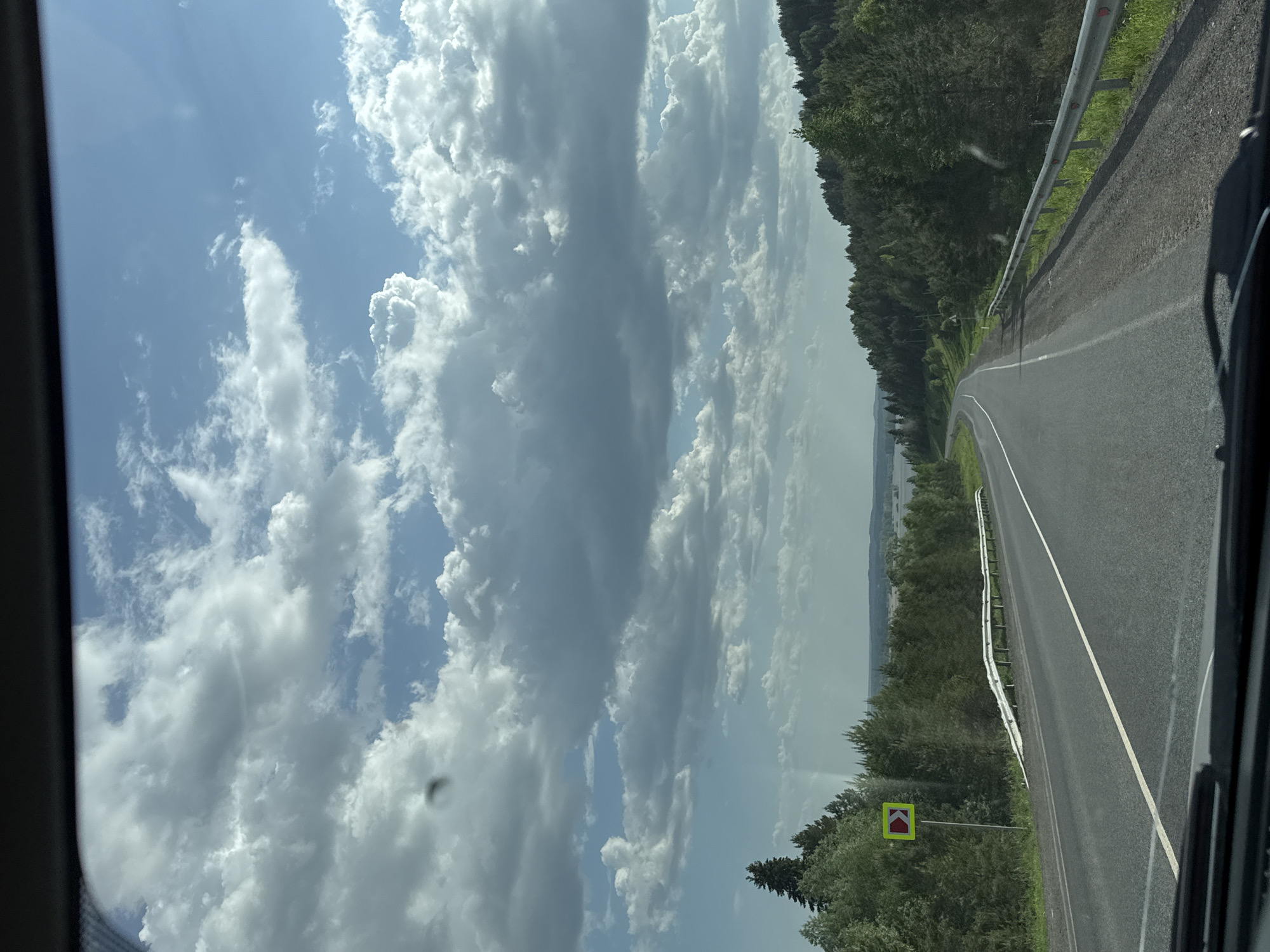 The image depicts a view from inside a vehicle traveling on a paved road near Ir'yak, Russia. The road curves gently to the right and has a white line dividing the lanes. A metal guardrail runs along the right side of the road, separating it from a forested area. A yellow and red traffic sign indicating a sharp curve is visible on the right side of the road. In the distance, beyond the trees, there appears to be a body of water. The sky is filled with numerous white and gray clouds. The viewpoint is from within a vehicle, evidenced by the vehicle frame on the left and bottom right of the image.
