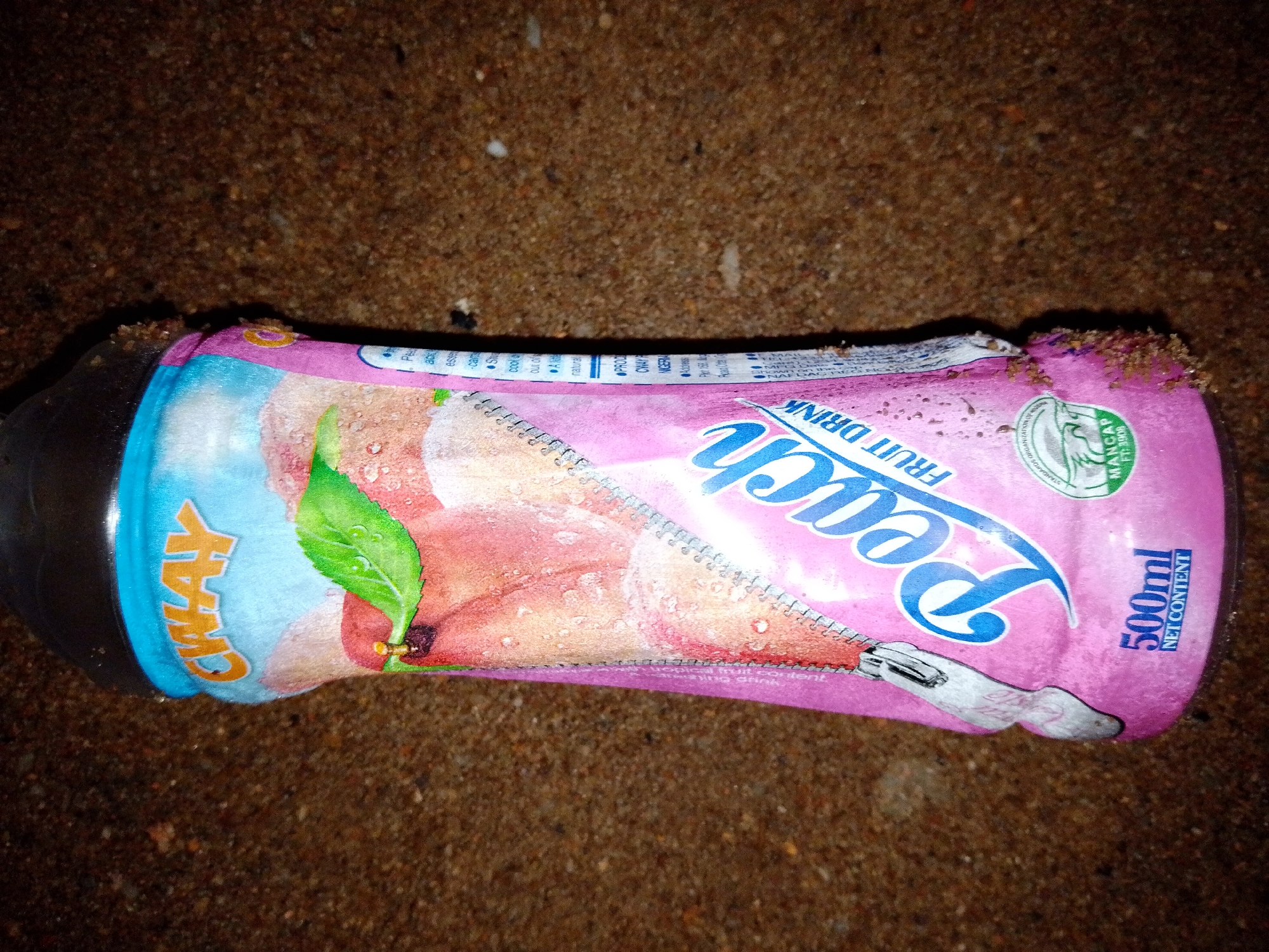 A pink and blue bottle of "Sway Peach Fruit Drink" is lying on sandy ground. The bottle has a blue cap and a label that features a peach, a green leaf, and a zipper graphic. The text "500ml NET CONTENT" is visible on the right side of the bottle. The ground appears to be a beach or a sandy area. The image is taken from above, looking down at the bottle. There are no people or animals visible in the image. The bottle appears to be slightly damp, with water droplets on its surface. The background consists of dark, sandy soil.