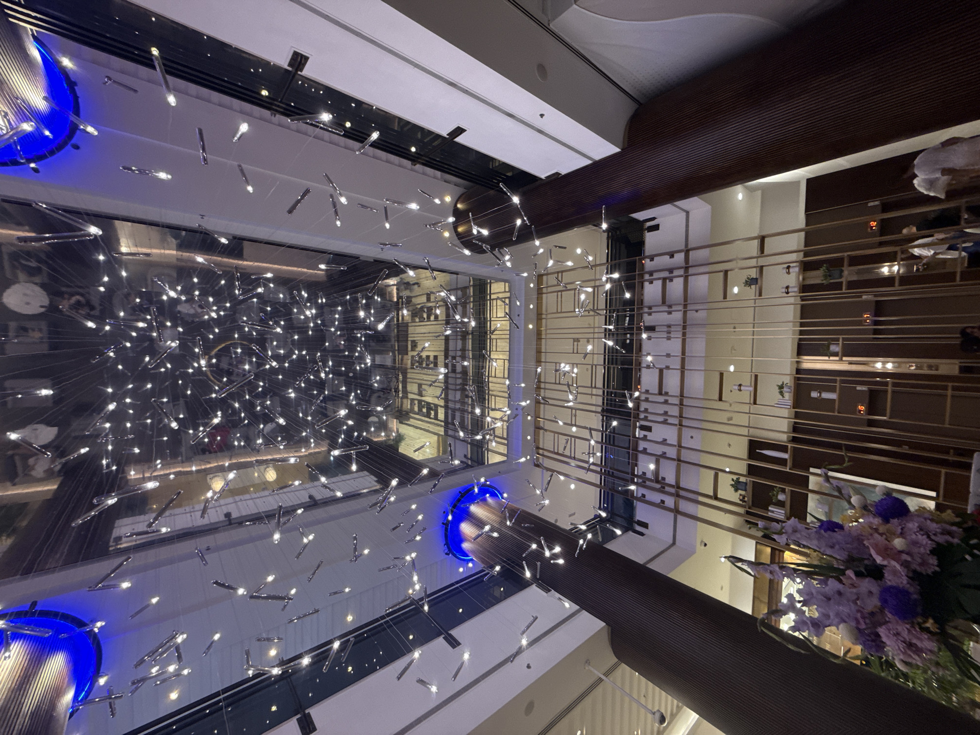 This is a photograph taken inside the Dubai Mall, United Arab Emirates. The camera is positioned looking upwards towards the ceiling, which features a striking overhead light installation. This installation consists of numerous small, elongated lights suspended from thin wires, creating a dazzling, star-like effect against the reflective ceiling.
Below the light installation, the atrium of the mall is visible, showcasing multiple levels with glass railings and balconies. Large, dark brown cylindrical columns, textured with vertical grooves, dominate the foreground and frame the view upwards. These columns appear to be structural elements supporting the mall's architecture.
Further down, a decorative wooden shelving unit is visible, featuring an arrangement of shelves that hold various items, including small potted plants and decorative vases. The shelves are outlined with vertical wooden beams, creating a geometric pattern.
In the lower portion of the image, a large floral arrangement with purple and pink flowers is positioned to the left. In the background, several individuals are visible walking on the lower levels of the mall, their figures somewhat blurred due to the distance and lighting. The overall ambiance suggests a modern, upscale shopping environment.