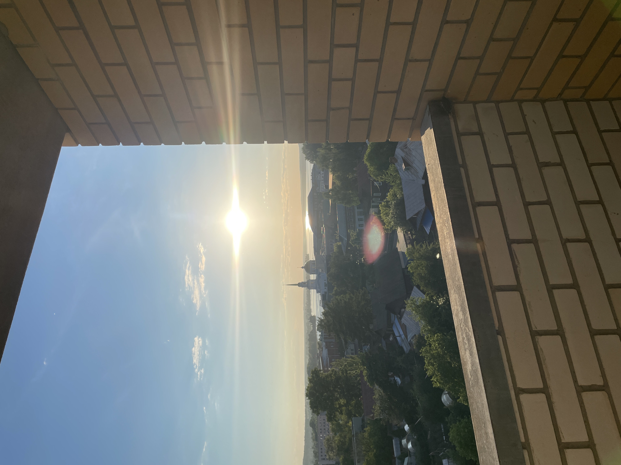 This high-angle urban landscape image captures the city of Votkinsk, Russia, during the golden hour, likely sunset. The viewpoint is from a building, with a light-colored brick wall and a dark concrete ledge forming the foreground and obscuring parts of the upper and right edges of the frame. The sun, positioned in the upper right, is low in the sky, emitting strong light and several lens flares. The sky is clear blue with a few sparse white clouds.
The mid-ground and background reveal a sprawling cityscape. Numerous buildings, predominantly residential with varied rooflines, are interspersed with abundant green deciduous trees. A significant body of water, identified as the Votkinsk Reservoir, is visible in the distance, reflecting the sunlight. On the far side of the reservoir, a prominent architectural structure, the Annunciation Cathedral of Votkinsk, stands out with its white facade, dark domes, and a tall, slender spire. No individuals are visible in the scene.