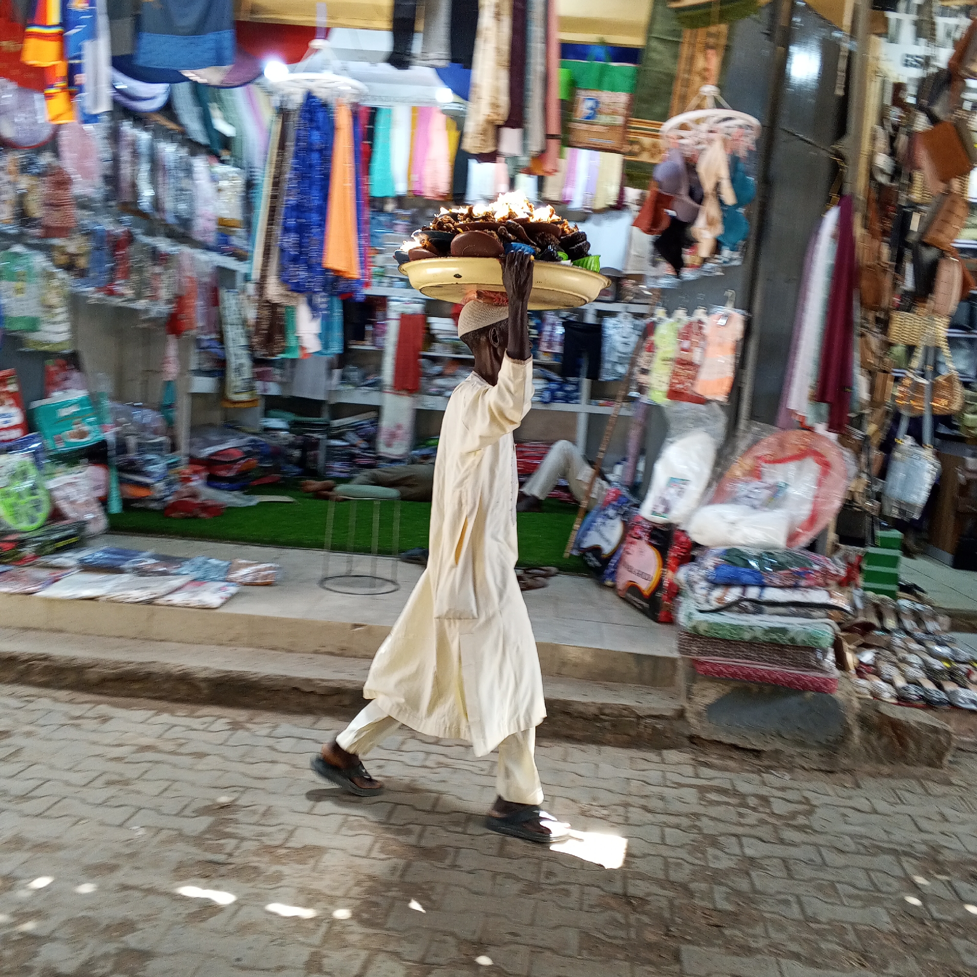 A man is walking on a cobblestone street carrying a large tray of food on his head. He is wearing a cream-colored robe and a white cap. The street is lined with shops selling various goods, including textiles, bags, and other items. The background shows a bustling market scene with stalls and more merchandise. The lighting suggests it is daytime. The man is in motion, indicated by the blur in the image. The location is Arawa, Nigeria.
