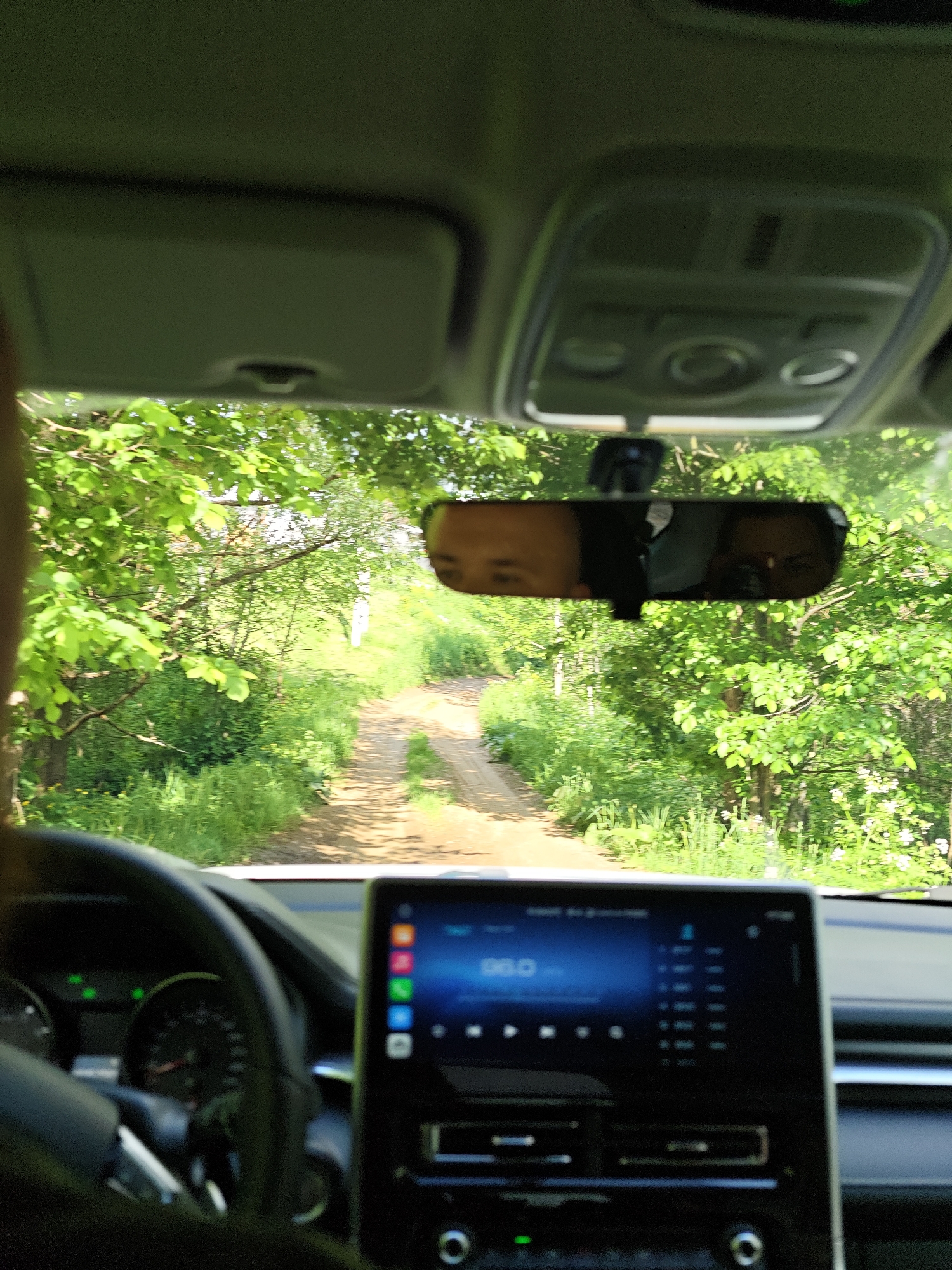 The view from inside a car driving on a dirt road lined with lush green trees. The rearview mirror reflects the faces of two individuals. The car's dashboard is visible, featuring a large infotainment screen displaying information. The steering wheel and part of the driver are also visible. The location is Zhigalki, Russia, as that was specified.