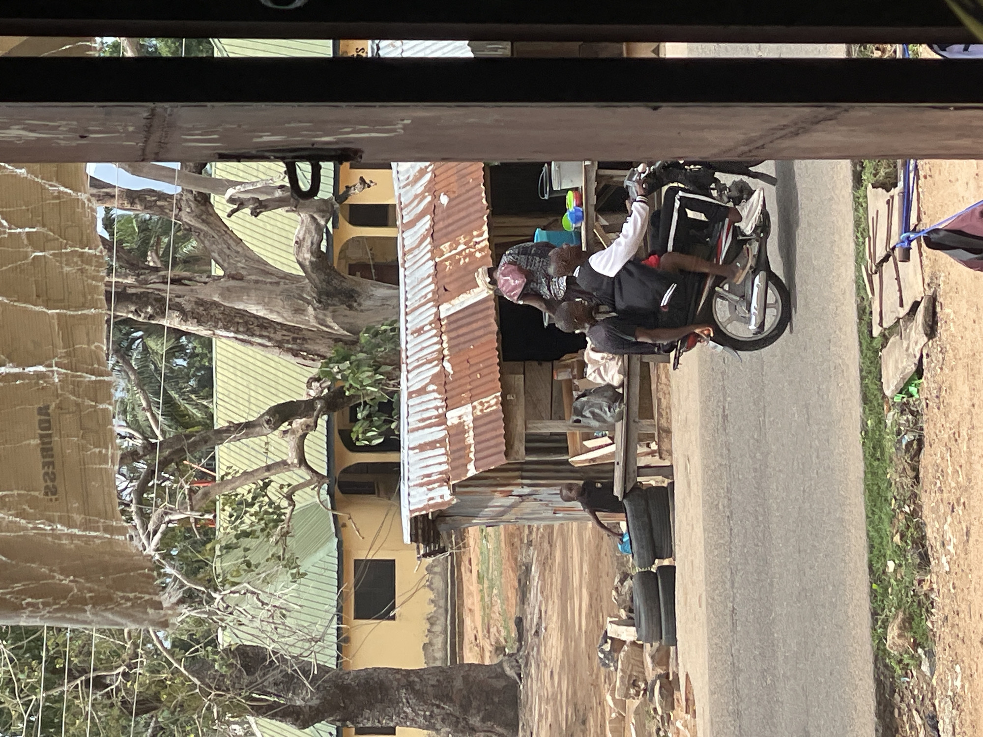 The image depicts an outdoor street scene, likely in Mararaba, Nigeria, under bright daylight. A paved road runs through the middle ground, separating the foreground from structures and activities. To the right of the road, the ground is dusty with patches of green grass.
Three individuals are visible. On the paved road, two people are seated on a silver and black motorcycle. The person in front wears a white and black jacket and dark pants, with a light-colored leg or foot resting near the ground. The second person, largely obscured, sits behind the first. To the left of the road, a person in a dark shirt stands near a rustic structure. This structure is characterized by a rusty corrugated metal roof and walls, surrounded by wooden planks and posts. Colorful plastic containers are visible near this structure, along with a stack of black tires.
In the background, there's a light yellow building with a green roof on the far left, and another yellow-walled building further back. Large trees with gnarled branches and some foliage are prominent on both sides of the scene and in the background.
On a surface in the far left foreground, partially obscured and mirrored, the word "ADDRESS" is faintly discernible. The scene captures an ordinary moment of daily life, with people on a motorcycle and an individual seemingly attending to goods or tasks near a roadside stall or establishment.