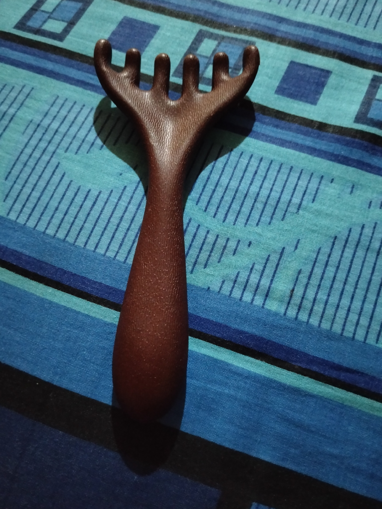 The image shows a brown hair comb with five prongs resting on a striped blue and black fabric. The fabric has alternating wide and narrow stripes. The comb is positioned diagonally across the fabric, with the prongs facing upwards and slightly to the left. The fabric appears to be a bedsheet or similar textile. The scene is lit from above and to the right, casting a shadow of the comb onto the fabric. The image is a close-up shot, with no visible background beyond the fabric and the comb. The location in Payagaboh, Indonesia, is not depicted in the image.