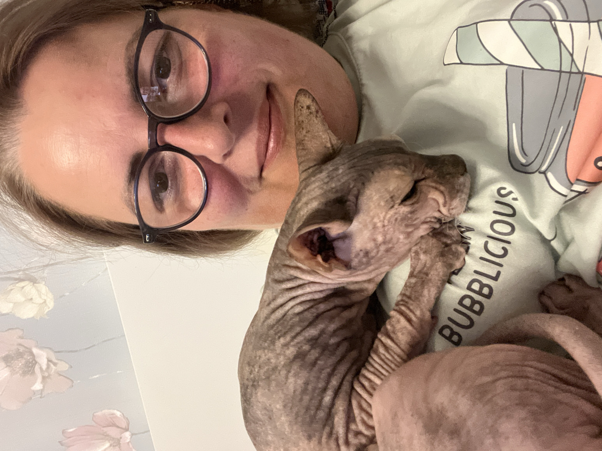 A close-up, selfie-style image shows a woman wearing glasses, holding a Sphynx cat. The woman is facing the camera, and the cat is nestled on her chest, with its head resting down. The cat appears to be sleeping. The woman's t-shirt has the word "BUBBLICIOUS" printed on it in black letters, along with a graphic of a stylized jar. The background wallpaper features a floral pattern with pink and white flowers. There are no discernible features to suggest the image was taken in the city of Korolyov, Russia.