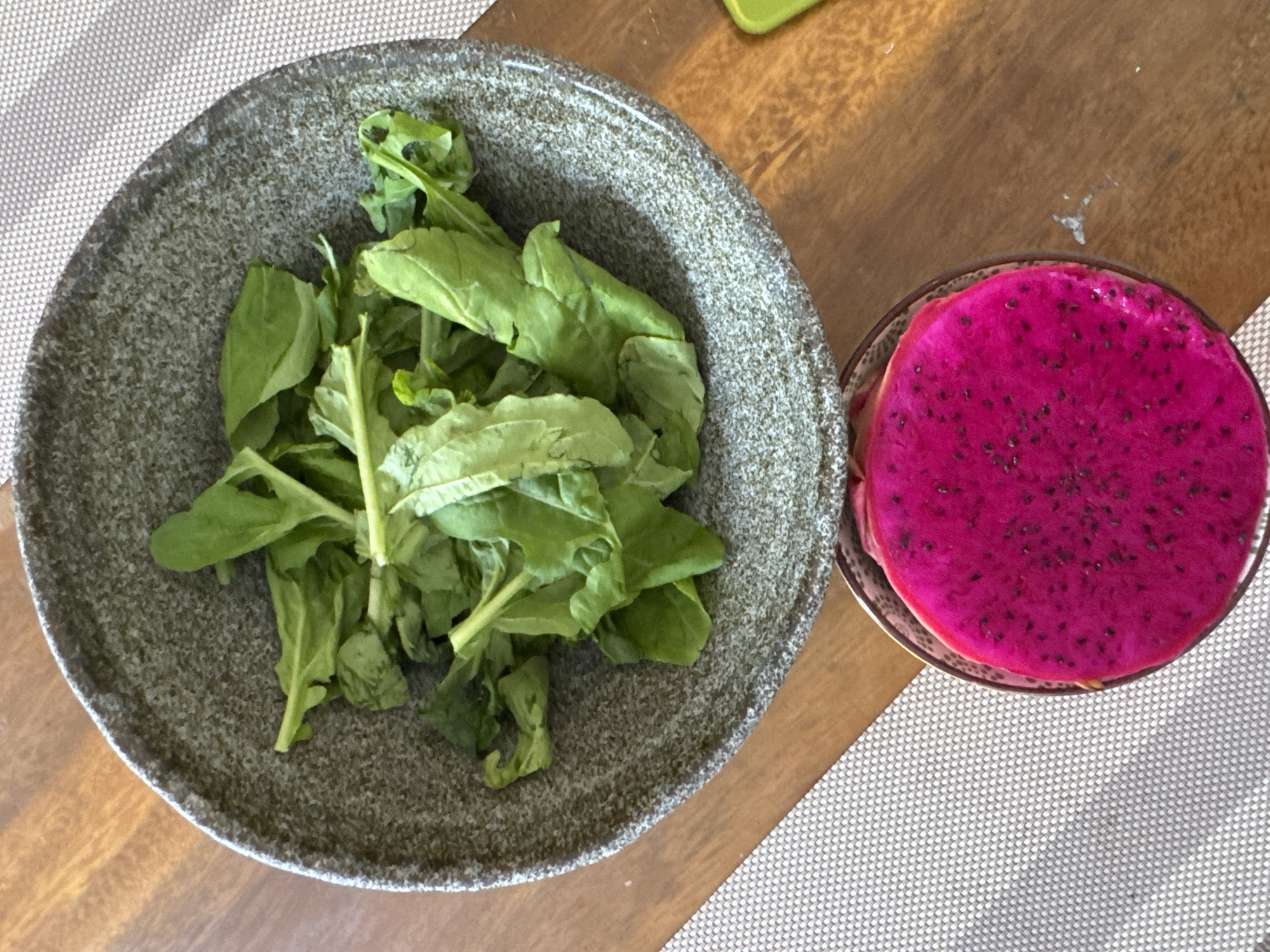 A top-down shot shows a light gray ceramic bowl filled with green spinach leaves next to a glass bowl with a bright magenta dragon fruit cut in half. Both bowls rest on a table covered with a textured, gray placemat, with the table's wood surface visible between them. The scene is illuminated by natural light, and the overall impression suggests a healthy meal. The location is not evident from the image, so I can't confirm the city of Kuta Selatan, Indonesia.
