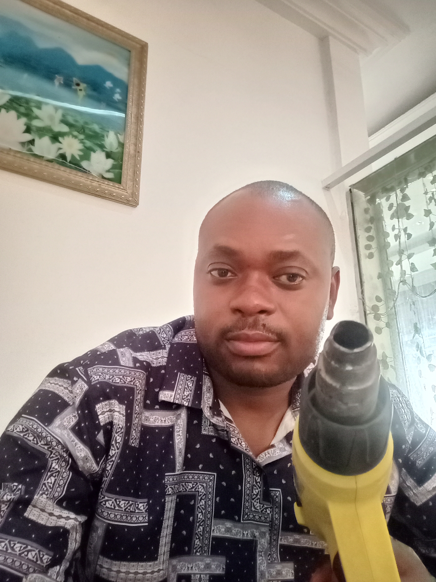 A dark-skinned man is holding a yellow heat gun in the foreground. He is wearing a dark patterned shirt with a white collar. The man is bald and has a slight mustache. In the background, there is a framed painting of a landscape with white flowers and a window with floral curtains. The location is possibly Gwarinpa, Nigeria, but there are no specific indicators in the image to confirm this.