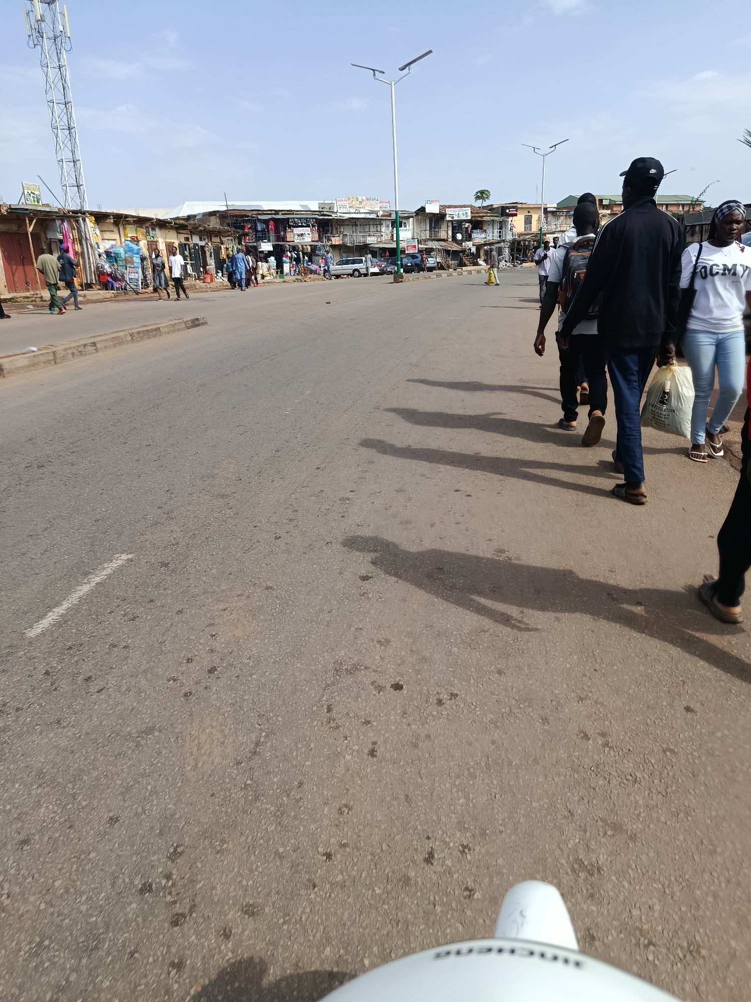 The image depicts a street scene in Jos, Nigeria, with a row of shops lining the right side of a paved road. Several people are walking along the road, mostly in the foreground and mid-ground. The shops appear to be open-air market stalls or small retail establishments. There are various goods displayed outside the shops, including clothing and other items. A cell tower is visible in the far left background. The sky is clear and blue, suggesting a sunny day. The road is asphalt and shows signs of wear, with some dark patches and a white line marking. Shadows of the people walking are cast on the road, indicating the direction of the sun. The overall impression is a busy, everyday street scene in an urban environment.