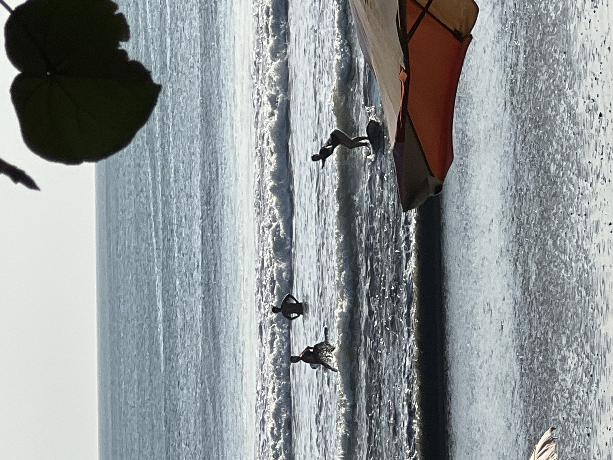 The image depicts a beach scene in Kuta, Indonesia, with several individuals engaged in water activities. In the foreground, a partially visible beach umbrella with orange and tan fabric shades a portion of the water. The ocean is choppy with white waves breaking. Several people are in the water. One person is on a surfboard, appearing to be actively surfing or balancing. Two other individuals are standing in the water, their bodies silhouetted against the bright sky. In the background, the horizon is visible where the ocean meets a pale sky. Dark green leaves hang from the upper right corner of the frame, casting a shadow and adding a natural element to the composition.