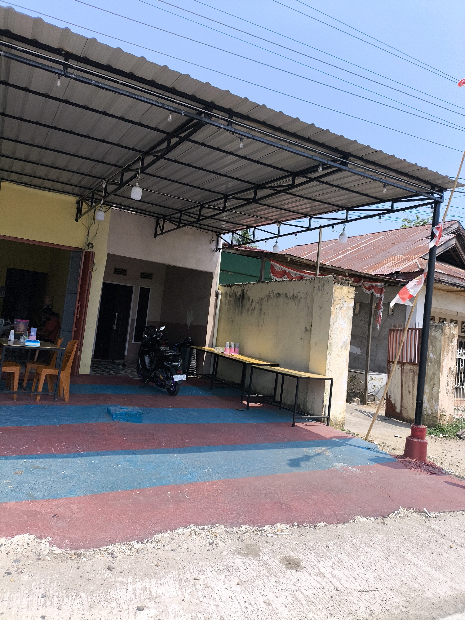 The image depicts an outdoor seating area of a food establishment, likely a warung or small restaurant, located in Kwandang, Indonesia. The foreground features a concrete ground painted with alternating red and blue stripes, showing signs of wear. To the left, a yellow building with an open entrance reveals a glimpse of an interior with tables and chairs. An orange plastic chair is visible in the seating area. In front of the yellow building, orange plastic chairs are arranged around a table.
Dominating the mid-ground is a black motorcycle parked on the red and blue striped ground. Behind the motorcycle, a building with a weathered, reddish-brown corrugated metal roof stands. This building has a textured, light-colored wall and an entrance. Above the seating area and extending across the mid-ground is a gray corrugated metal roof supported by a black metal frame. Several light bulbs hang from the roof structure.
Further to the right, a long, light-colored wooden table with black metal legs is positioned outdoors. On this table, several pink and white cups are arranged. A tall black pole with a red base stands to the right of the wooden table, adorned with a red and white Indonesian flag. The background shows more buildings with corrugated metal roofs, vegetation, and power lines against a clear blue sky. The overall scene suggests a casual, open-air dining environment in a tropical climate.