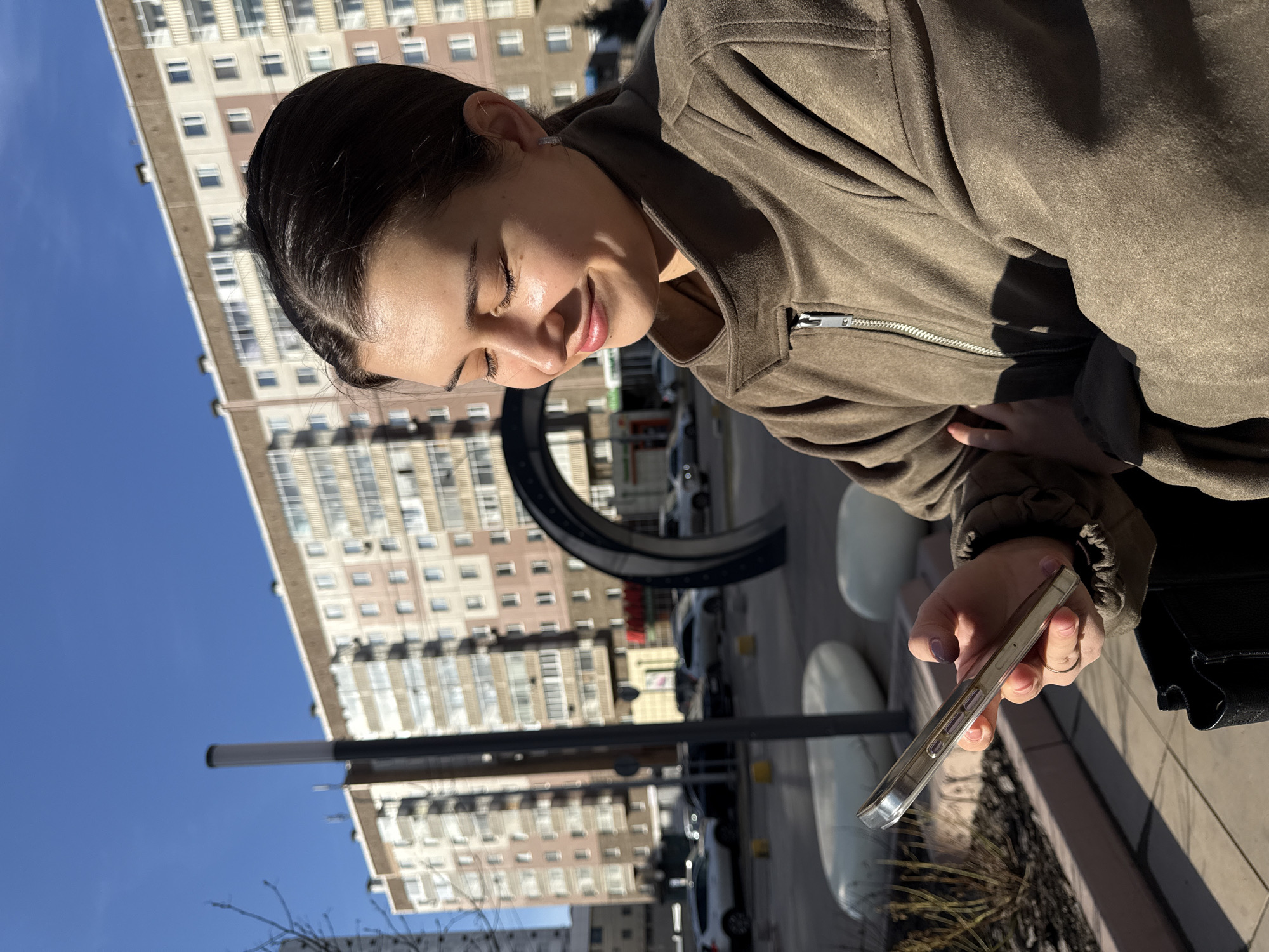 The image captures a young woman outdoors in an urban setting in Krasnoyarsk, Russia, under clear blue skies. The woman, with dark hair pulled back, is seen from the chest up, smiling with her eyes closed, as if basking in sunlight or reacting to something on her phone. She wears an olive-green or brown zippered jacket. In her right hand, she holds a smartphone in a clear case, her fingers, with dark nail polish, wrapped around it. A small, sparkling earring is visible in her left ear.
The background features a tall, light-colored apartment building with numerous windows, indicative of a city environment. Below the building, a street scene unfolds with parked cars and what appear to be white, rounded street fixtures, possibly benches or planters. A large, dark, circular art installation or sculptural element is partially visible in the middle ground. A slender, dark pole also extends upwards on the left side of the frame.
The bright sunlight and distinct shadows suggest it is daytime, likely morning or afternoon, with pleasant, clear weather. The woman's closed eyes and smile convey emotions of contentment, happiness, or amusement. There is no visible text in the image.