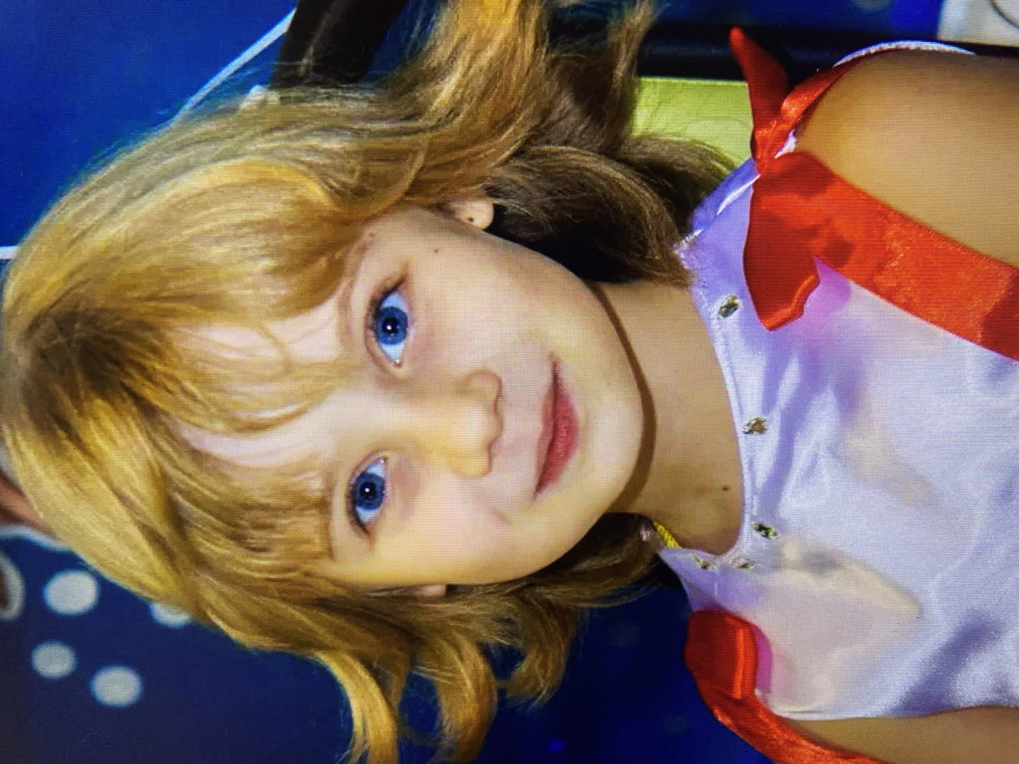 The image is a close-up portrait of a young girl with blonde, wavy hair and bright blue eyes. She is wearing a light purple dress with red bows on the shoulders and a decorative neckline. The background is dark blue with white circular patterns. The girl is looking directly at the camera and smiling. The image was taken in Stavropol, Russia.