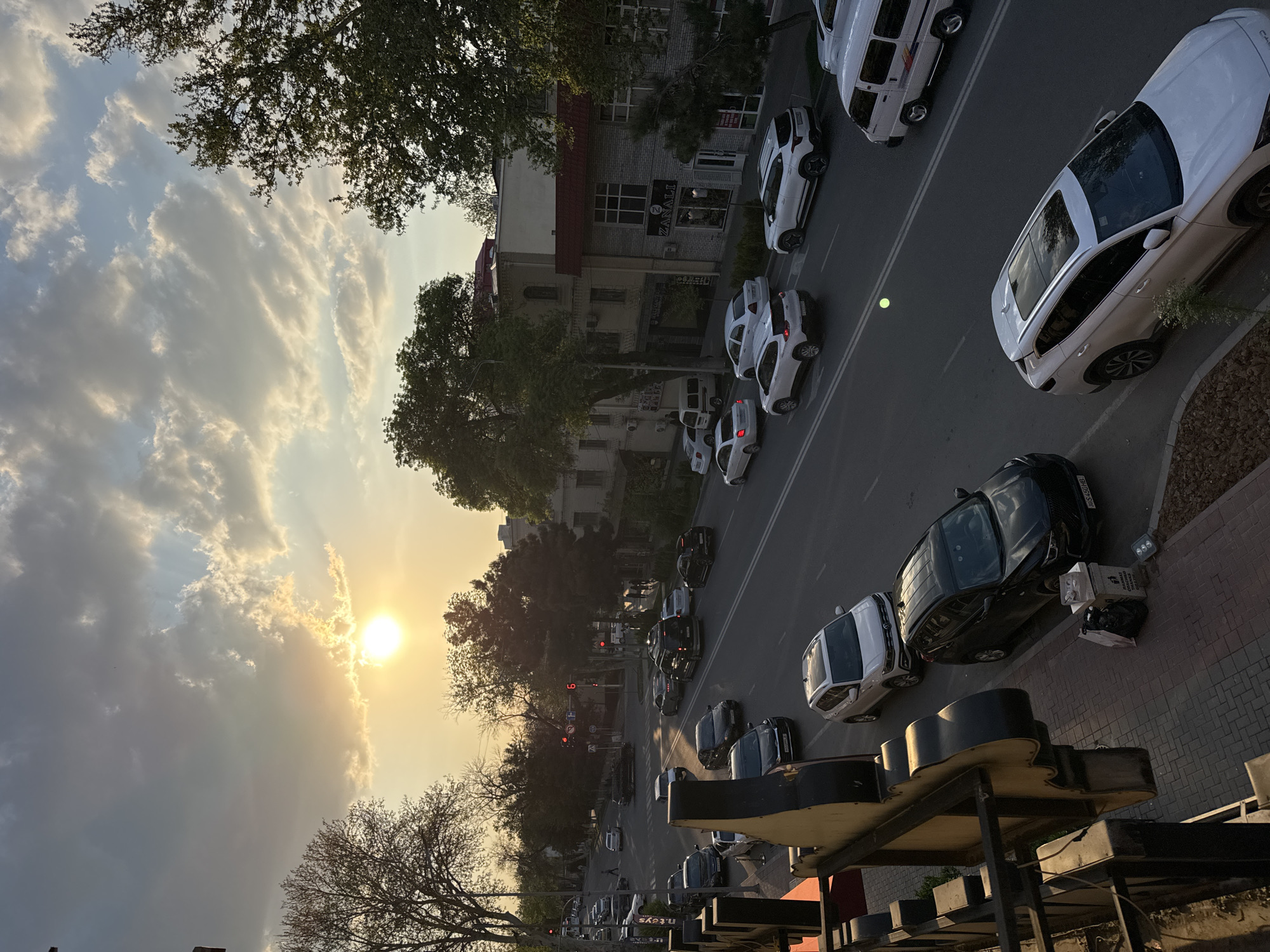 The image shows an elevated view of a street in Samarkand City, Uzbekistan at sunset. The street is lined with buildings and trees, and cars are parked on both sides. A line of primarily white cars are parked on the right side of the street, closer to the camera's perspective. Black cars are visible on the other side of the street in the lower part of the image, parked alongside lighter cars that are harder to discern due to the distance. The sun is low in the sky, casting a warm glow over the scene. The sky is filled with clouds, and the sun is partially obscured by the clouds. The presence of a sign with a stylized design in the foreground suggests the image was taken from a commercial or residential building. A building further up the street appears to house at least one business, indicated by visible signage.