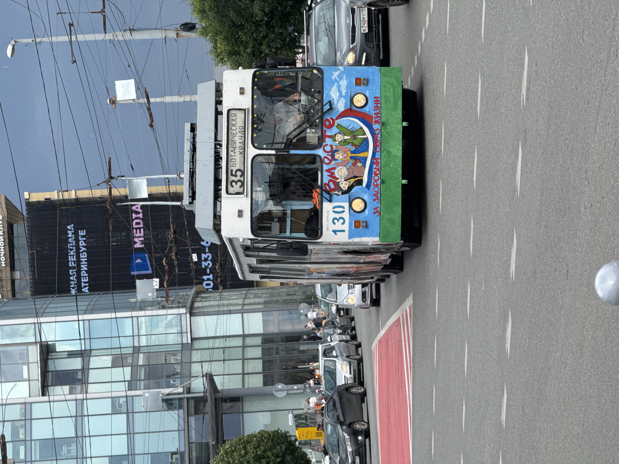 In Yekaterinburg, Russia, a trolleybus with the number 35 and the destination "Botanicheskaya - Kratsya" is captured on a street. The trolleybus is decorated with a mural depicting cartoon characters and the slogan "For a healthy lifestyle" in Russian. It is in motion, alongside other vehicles, including cars. The scene appears to be a typical urban setting with buildings and overhead wires visible. The sky is overcast, suggesting possibly rainy weather.