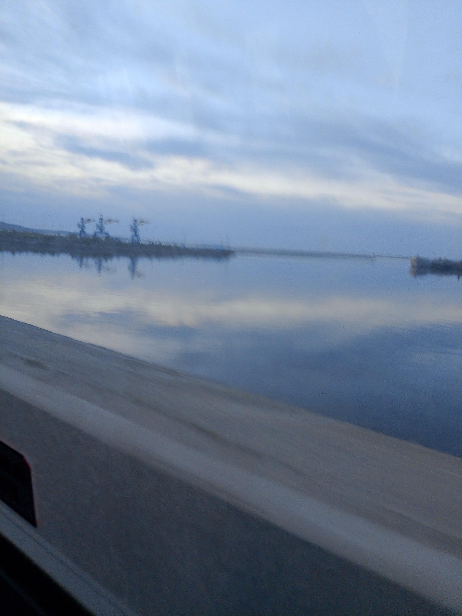 The image shows a waterfront scene in Chaikovsky, Russia. In the distance, structures resembling cranes are visible along the shoreline. The water reflects the cloudy sky above. In the foreground, a gray, linear structure, possibly a railing or a part of a vehicle, is blurred, suggesting movement. The overall environment appears to be a body of water, likely a river or reservoir, under an overcast sky.
