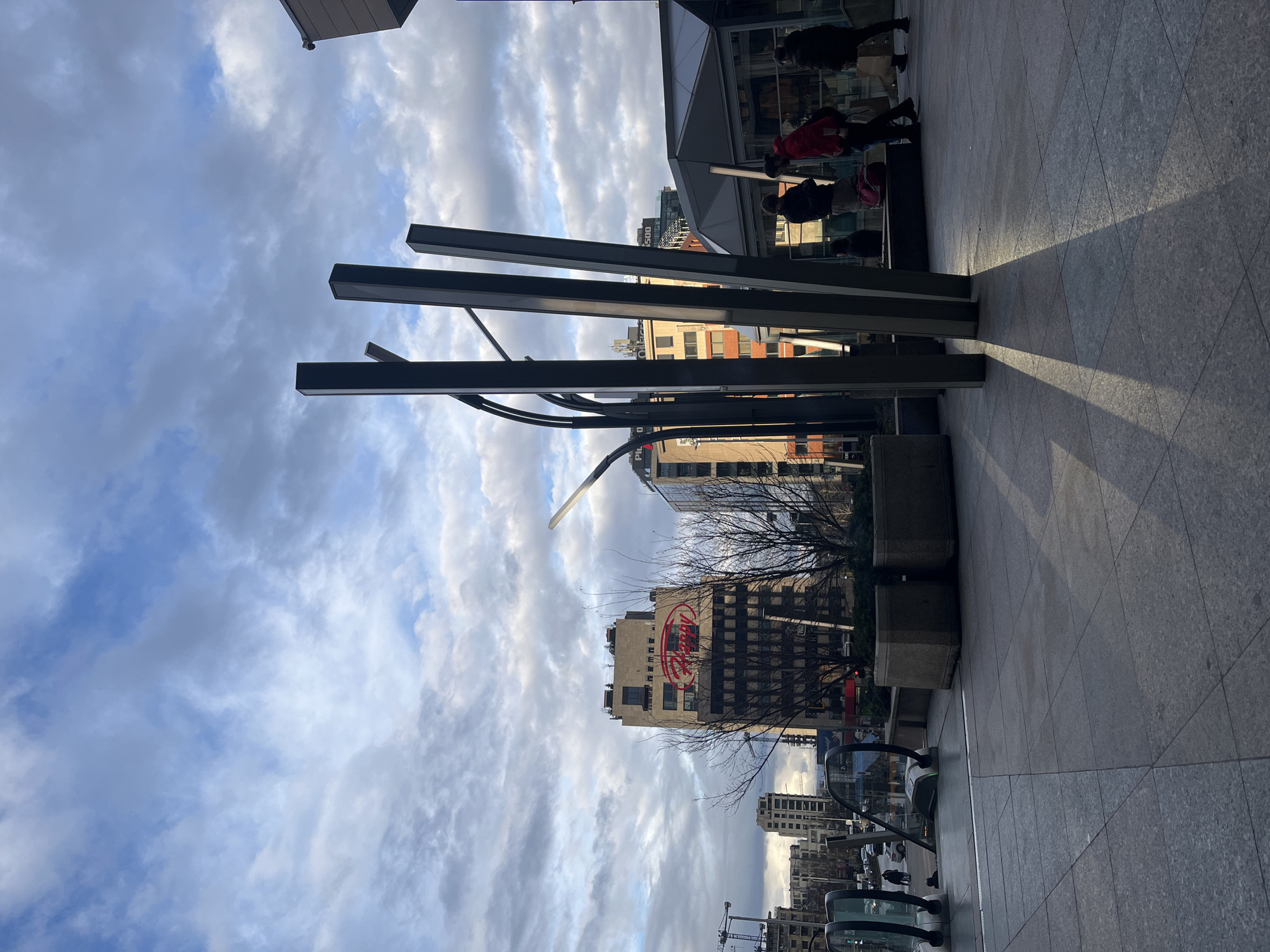 Captured from ground level, the image showcases an urban scene under a cloudy sky, with buildings and city infrastructure dominating the view.
The foreground features what appears to be a paved walkway or plaza, reflecting sunlight. Vertical metal architectural elements stand prominently, casting shadows on the ground.
In the middle ground, a cluster of buildings is visible, including one with a large red "Nestlé" logo, identifying the location as Sofia, Bulgaria. Bare tree branches frame the buildings.
Vehicles line the street, including what seem to be electric scooters. People are visible near a building's entrance.
The background consists primarily of a cloudy sky, providing an atmospheric backdrop to the urban landscape.
