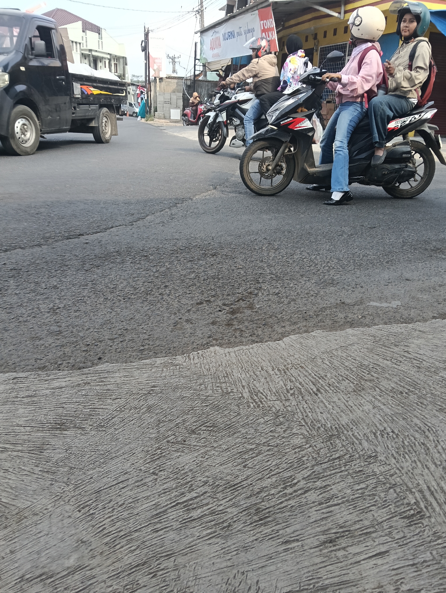 In Puspahiang, Indonesia, the image captures a street scene with several motorbikes. In the foreground, a black pickup truck is parked on the left side of the road. The road itself is asphalt. A motorbike with two female occupants is prominently featured in the center, with the driver wearing a light pink shirt and blue jeans, and the passenger in a beige jacket and dark pants. Both are wearing helmets. Another motorbike with two riders, also wearing helmets, is visible behind them. A third motorbike is further back, partially obscured. Buildings line the street in the background. The sky appears overcast.