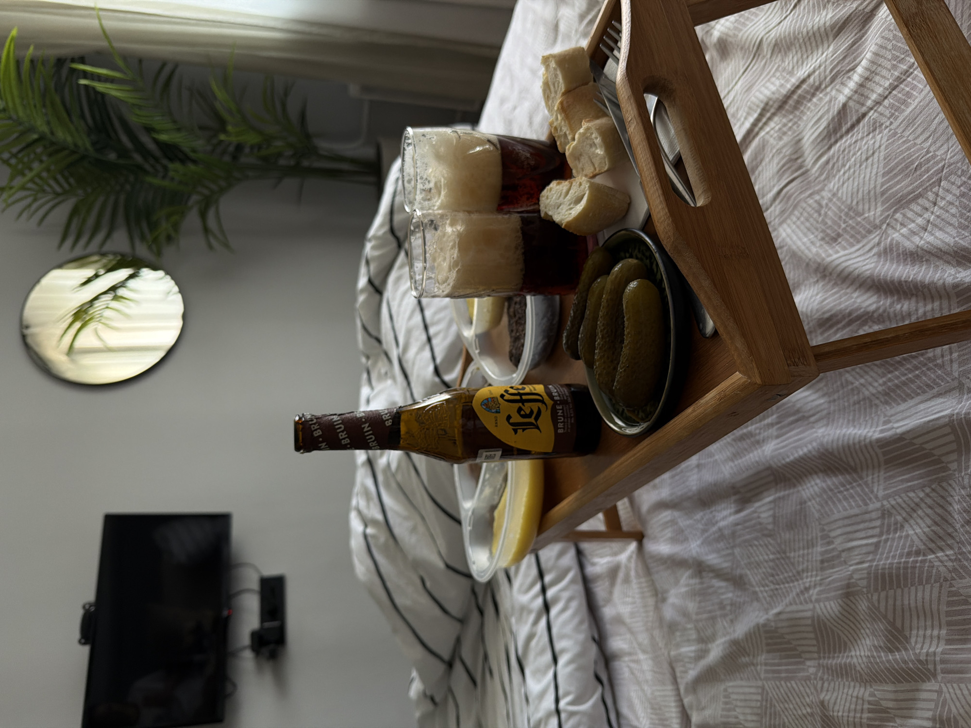 A still life presents a wooden tray perched on a bed covered with a striped and geometric patterned bedspread. The tray holds a variety of items: two partially filled glasses of beer, chunks of bread, a bowl of pickles, a small container of a yellow substance, and a bottle of beer labeled "Leffe Brune". A fork and knife are also on the tray. To the left of the tray is a potted plant and a round mirror on the wall. Further to the left is a television mounted on the wall. The location is not discernible as being in Khimki, Russia based on the image alone. There are no people visible in the image.