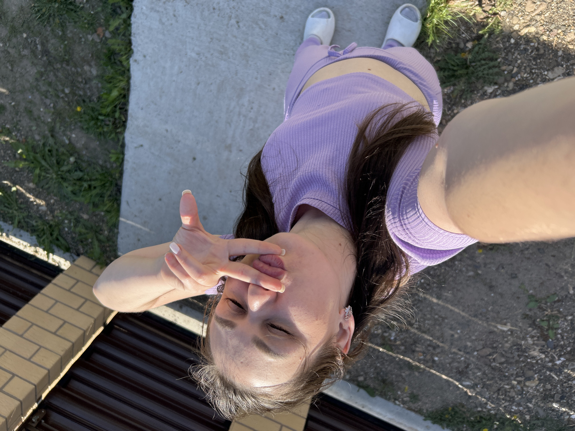A high-angle selfie shows a woman in a purple tracksuit and white sandals standing on a concrete surface next to a dirt area with sparse grass. She is making a face with her eyes closed and her tongue sticking out, holding her fingers near her mouth. In the background, there is a brick and dark brown metal structure, and an area with dirt and sparse grass. The photo was taken outdoors during daylight hours, casting shadows on the ground. The location is Chaikovsky, Russia.