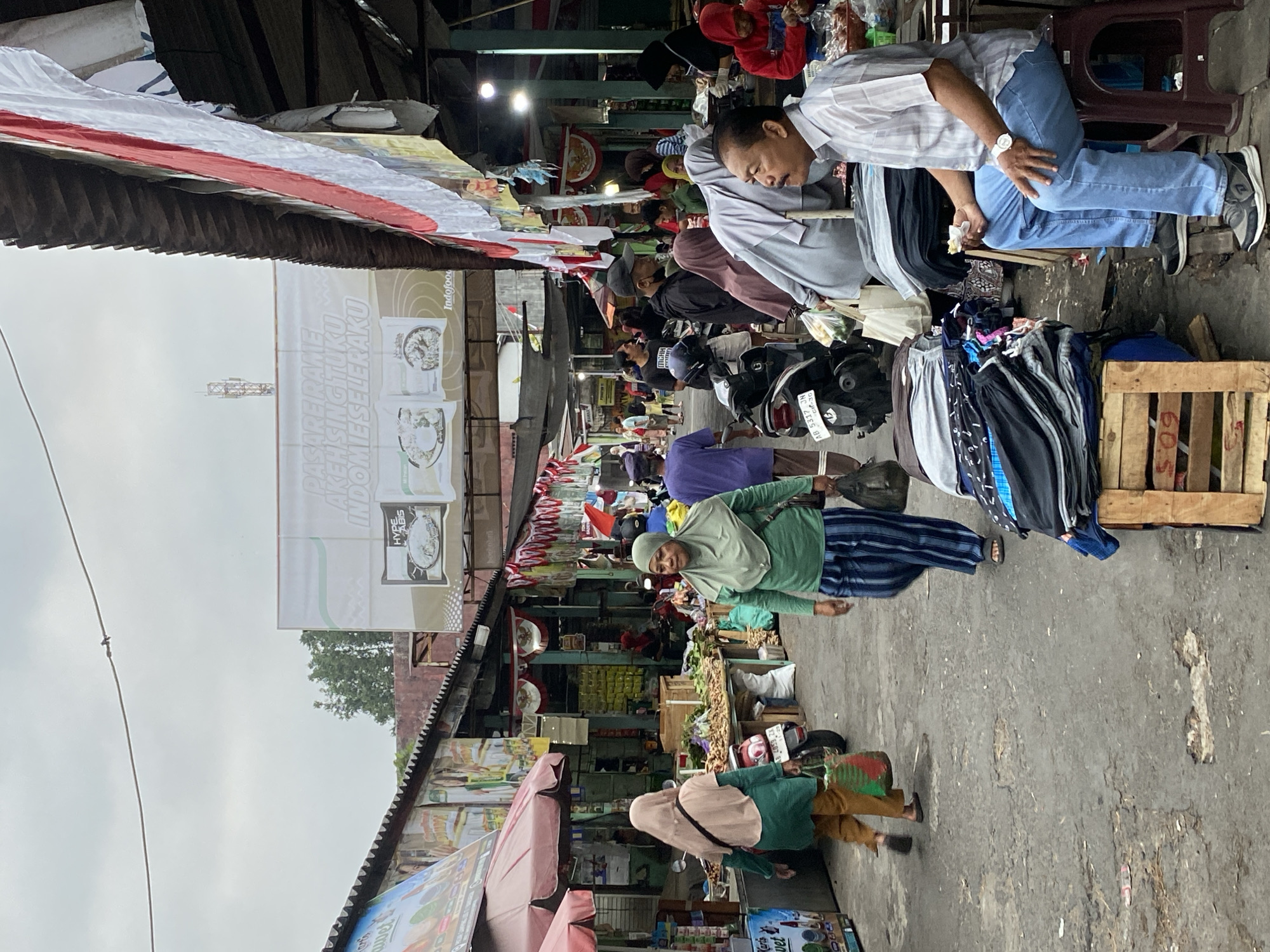 The image depicts a bustling marketplace in Ambarketawang, Indonesia, likely a scene on a cloudy day given the ambient lighting. The market is characterized by rows of stalls with corrugated metal roofs and awnings, decorated with red and white Indonesian flags. The ground is a worn, paved surface with visible cracks and debris.
In the foreground, a man is seated on a dark plastic stool to the right, wearing a striped light-colored short-sleeved shirt and blue jeans. He is holding a small white item, possibly food, in his right hand and looking to his left. In front of him, stacked neatly on wooden crates, are piles of folded clothing, primarily pants in various colors and patterns.
To the left of the seated man, and further back in the scene, a woman wearing a light green hijab and a striped blue and white outfit is walking towards the right. She is carrying a dark bag in her right hand. Another woman, wearing a peach-colored hijab and a teal-colored long-sleeved shirt over orange pants, is walking towards the left. She is carrying a patterned shopping bag.
Various other people are visible in the midground, some walking, some interacting at stalls. Motorcycles are parked along the pathways. Stalls display a variety of goods, including what appears to be fresh produce on wooden tables. A prominent banner hangs overhead, featuring images of food products and text in Indonesian. A telecommunications tower is visible in the far distance against the sky. The overall impression is one of a vibrant and active local market.