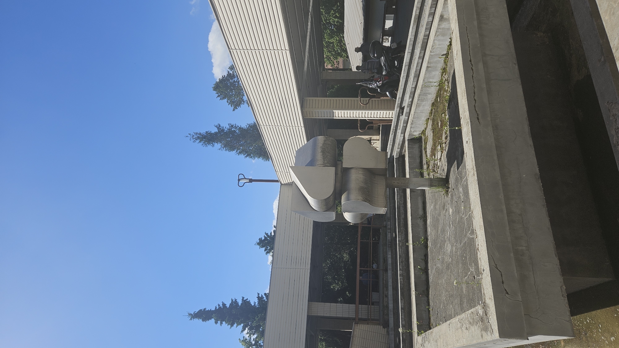 The image depicts an outdoor scene with a modernist sculpture in the foreground, set within a concrete structure that appears to be a dry fountain or water feature. The sculpture is made of metallic material and has an abstract, geometric form. Behind the sculpture, a building with a white, horizontally paneled facade and pillars is visible, suggesting a public or commercial space. There are also trees and a clear blue sky with a few clouds. In the middle ground, two men are visible near a motorcycle parked under the building's overhang. The overall environment appears somewhat aged, with some cracks in the concrete and sparse vegetation within the fountain area. The context of Prešov, Slovakia, is not visually ascertainable from the image itself.