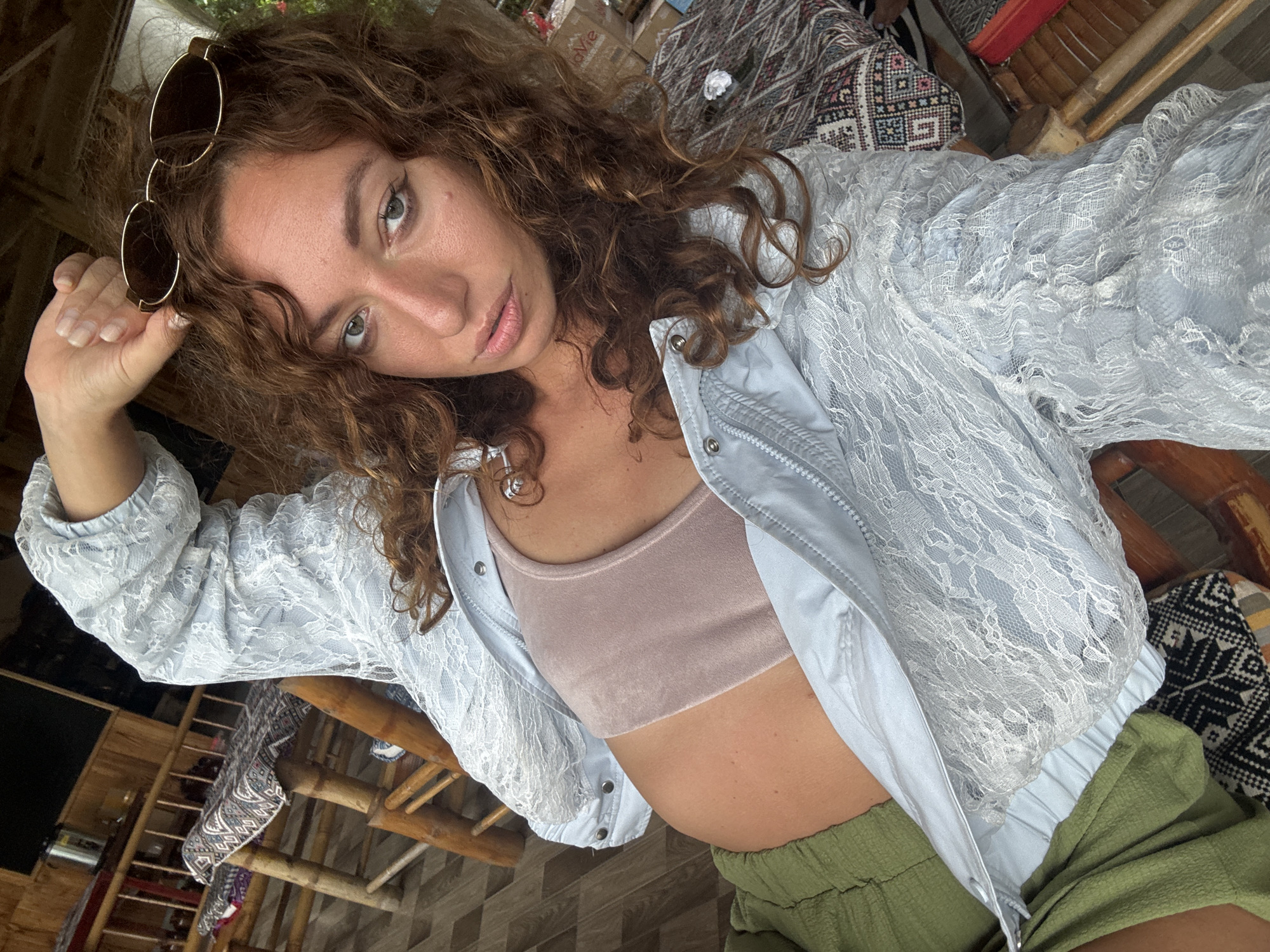 A young adult woman with curly brown hair, wearing a pale blue lace jacket, a pink crop top, and green shorts, is taking a selfie in an outdoor setting. She is resting her right arm above her head, holding a pair of sunglasses. The background suggests a rustic environment, with bamboo furniture and patterned textiles, indicative of a location such as Xóm Khán, Vietnam. The woman's expression is relaxed, and the overall tone of the image is casual and personal.