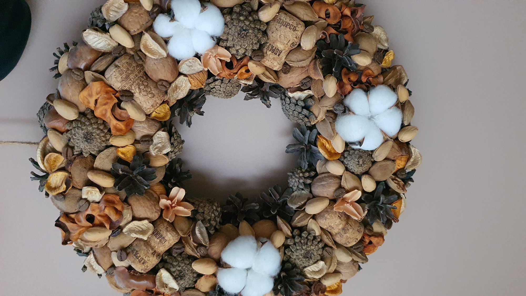 The image displays a circular wreath composed of natural dried materials such as pinecones, nuts, coffee beans, and dried flowers. Three white cotton-like accents are visible, one at the bottom left, one at the top right, and one slightly to the left of the center. The background is a plain, light beige wall. There is no indication of the city of Krasnoyarsk, Russia, or any urban environment within the image.