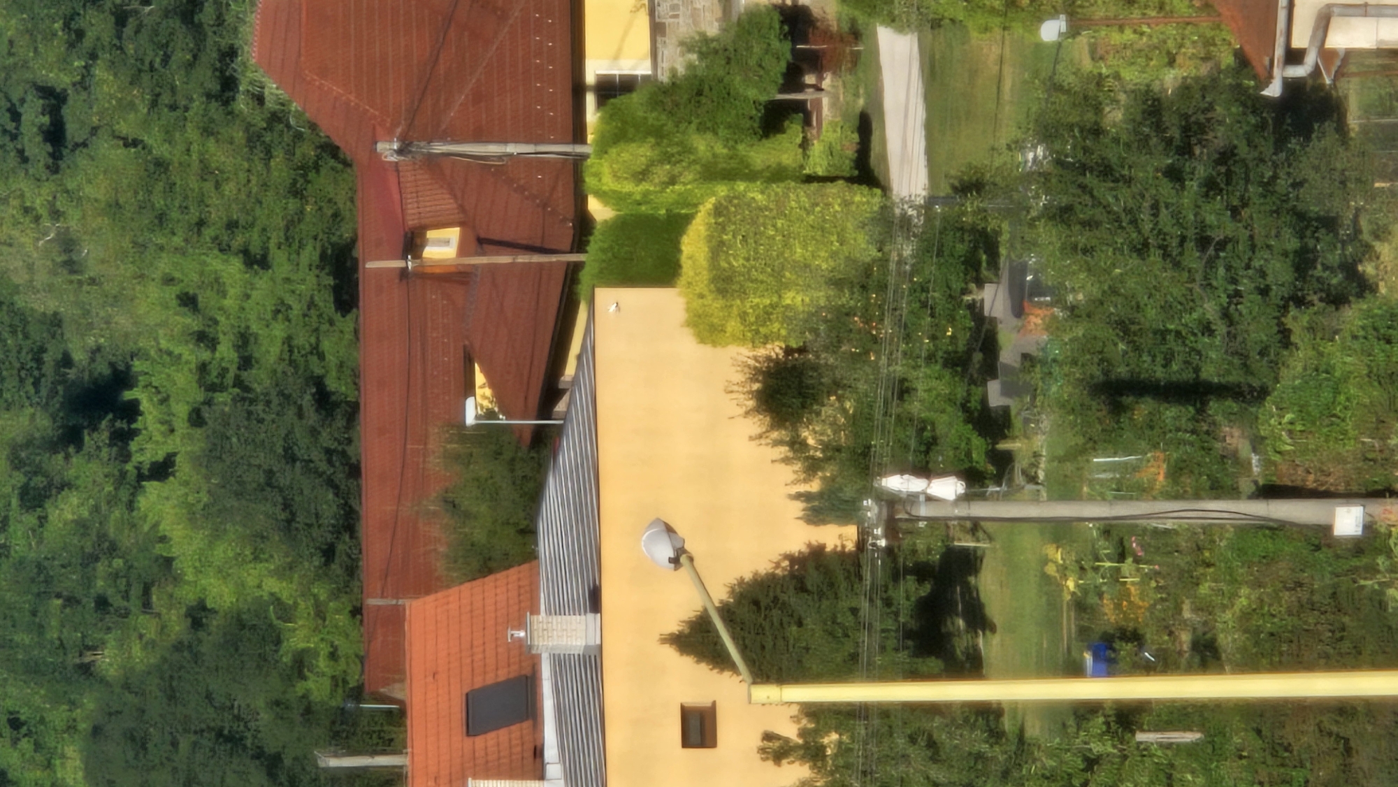 This is a medium-distance, daylight, outdoor, vertical shot of a residential area in Prešov, Slovakia. The foreground features a light yellow building with a small dark rectangular window. A tall, thin, light yellow pole with a street lamp extends upwards from the left side of the frame, passing in front of the building. Behind the building and to the right, there are manicured green bushes and trees. Beyond this, a darker, larger tree is visible. A grey utility pole with wires and a white object attached to its top stands to the right of the yellow pole. In the background, several houses with terracotta-tiled roofs are visible, nestled amongst lush green trees. The overall scene depicts a tranquil suburban landscape with architectural elements integrated into a verdant environment.