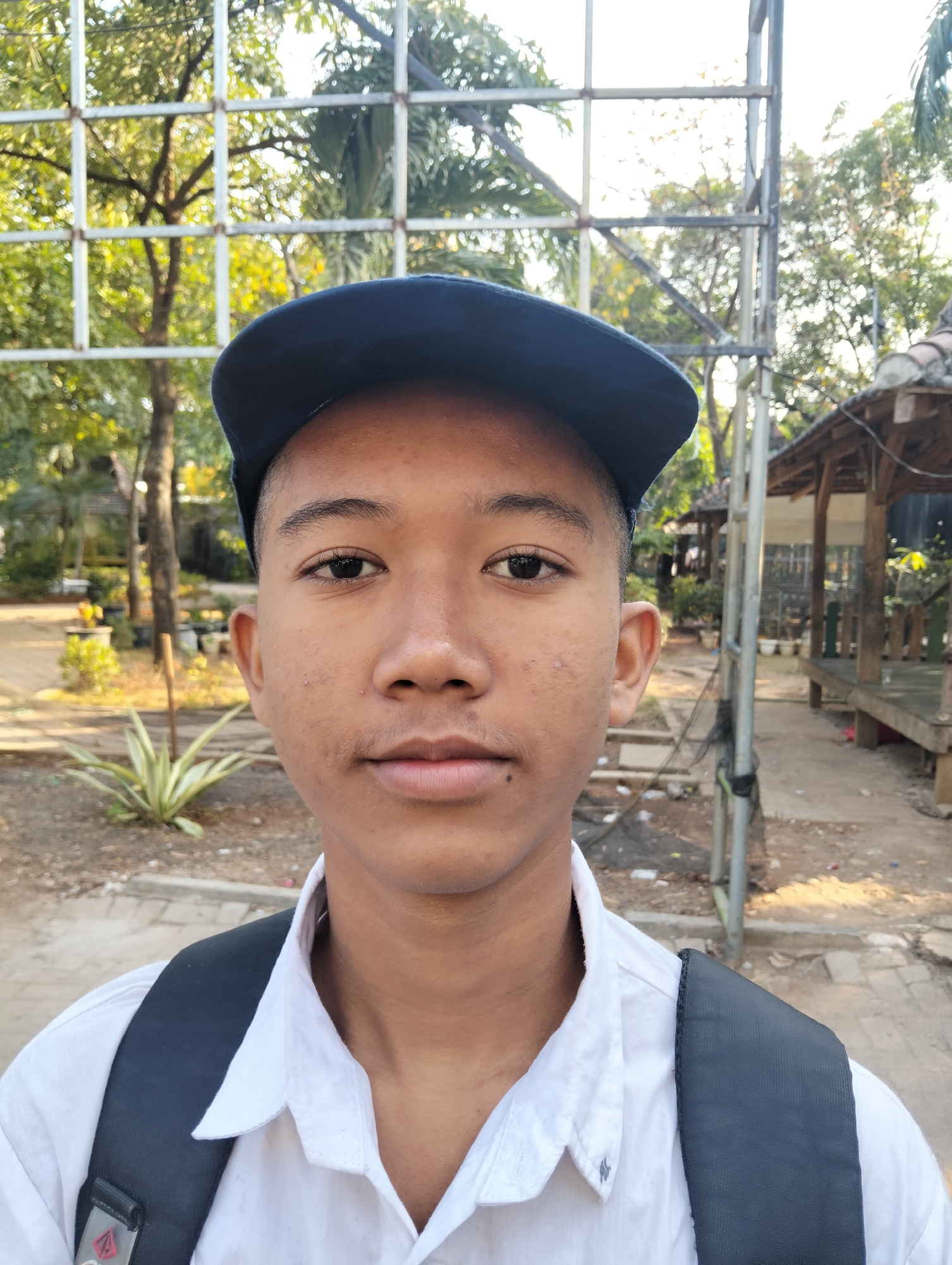 The image is a medium shot portrait of a young male wearing a blue cap, a white collared shirt, and a dark backpack. He is looking directly at the camera with a neutral expression. The background appears to be an outdoor environment with trees, a fence, and a wooden structure, possibly in a school or public area. The lighting suggests it is daytime. The location is identified as Paciran, Indonesia.