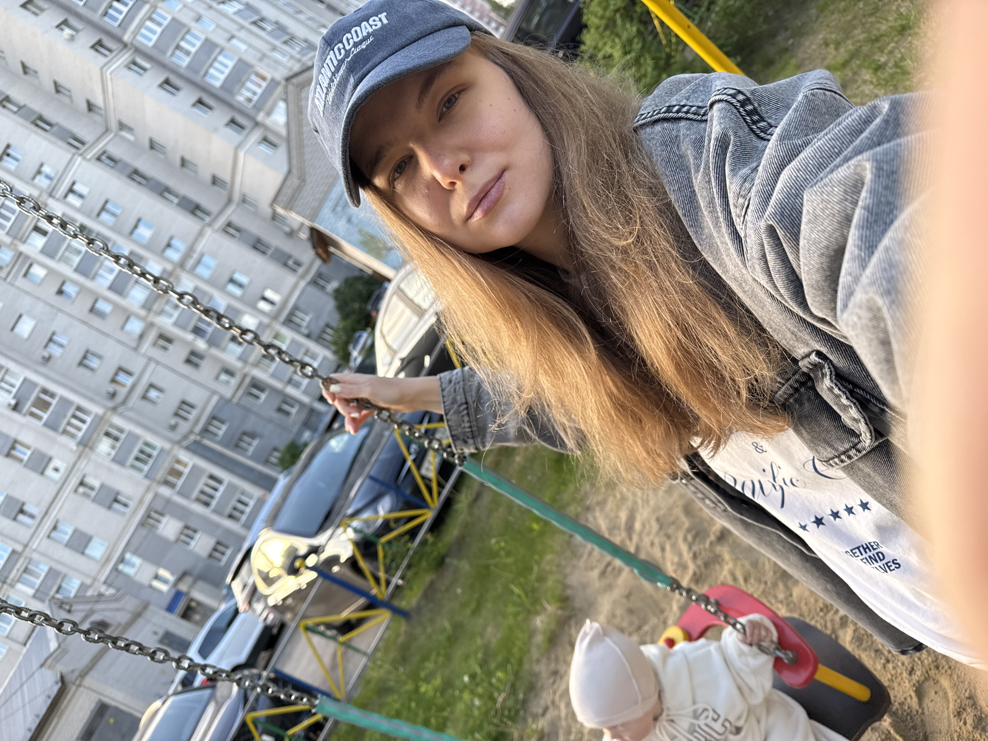 A woman wearing a baseball cap and a denim jacket is taking a selfie on a playground in Vladimir, Russia. She is holding a swing with a child in it. In the background, there is a tall apartment building, parked cars, and green grass.
