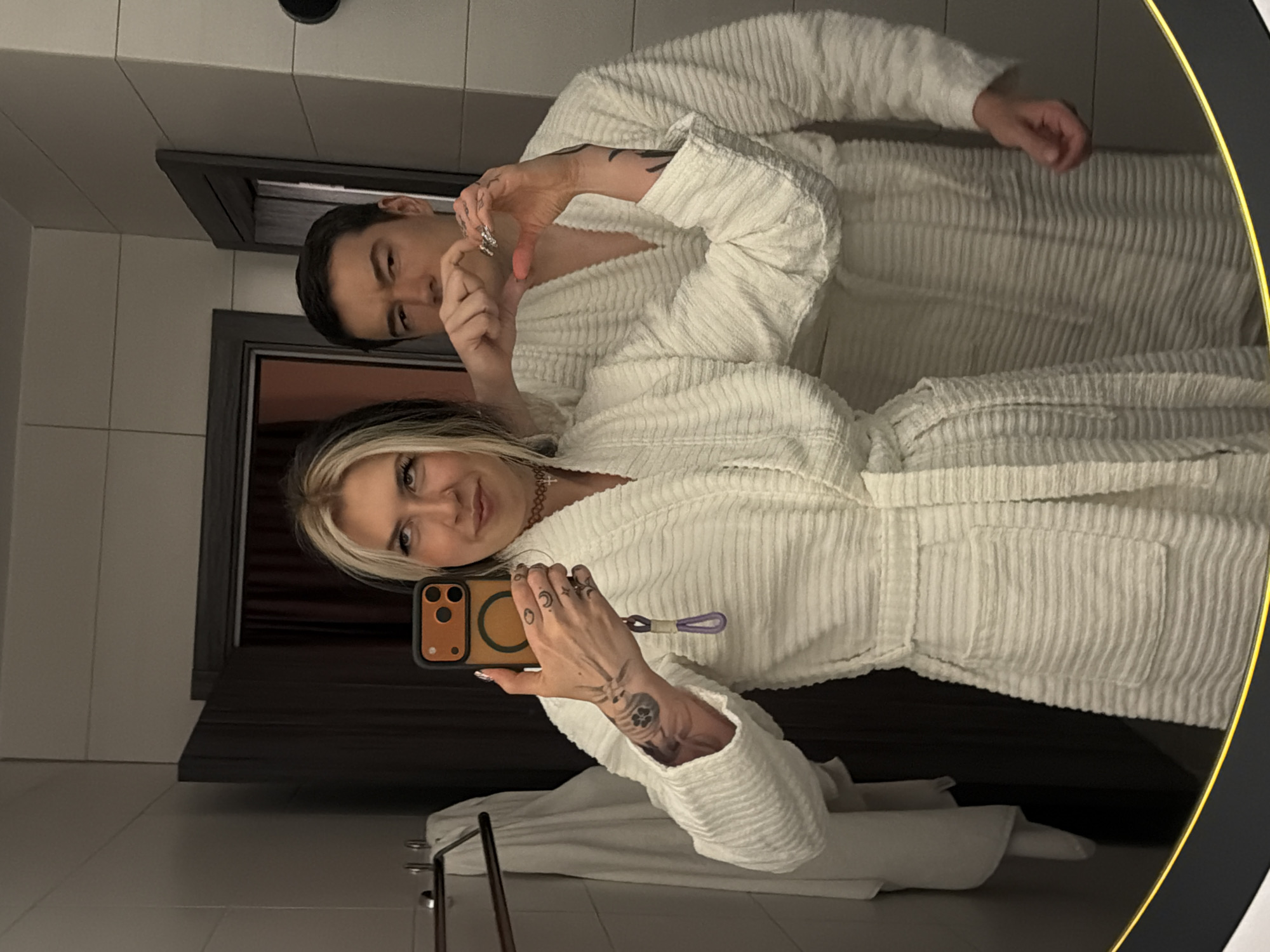 A man and a woman in white bathrobes are taking a selfie in a bathroom. The woman is holding the phone, and they are both making a heart shape with their hands. A mirror surrounds the scene. Behind them, part of the bathroom is visible, featuring a dark curtain. The location is likely a hotel or spa within Palilula Urban Municipality, Serbia.