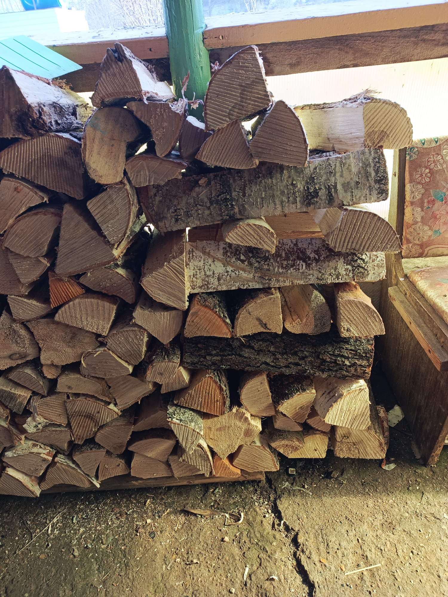 The image shows a stack of firewood. The wood is arranged in a pile against a wall. Some of the pieces have bark, and some are split. The ground is dirt and wood chips. The location and city cannot be determined from the image alone.