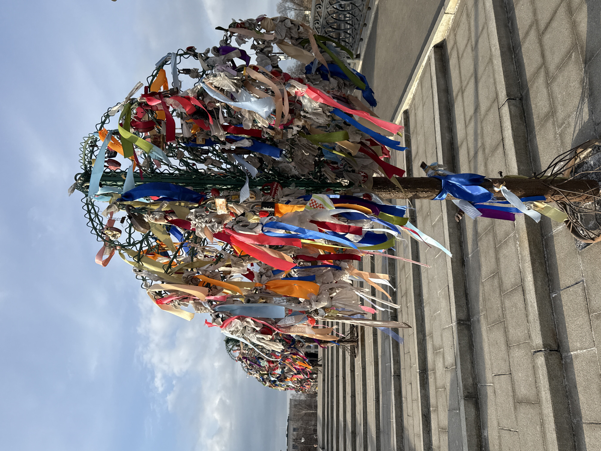 A fabricated metal "love tree" stands prominently in the foreground, constructed with a central brown-painted trunk and an intricate, rounded green metal wire canopy. This structure is densely adorned with hundreds of multi-colored fabric ribbons, including shades of red, blue, yellow, green, pink, orange, and white, many appearing weathered. Numerous metallic padlocks, both heart-shaped and rectangular, are also secured to the wire branches. The tree rests on a pedestrian area composed of grey rectangular paving stones, arranged as a series of shallow, broad steps. To the left, a linear arrangement of identical, similarly adorned metal love trees recedes into the distance. On the right, a dark, ornate metal railing borders the paved walkway, indicating an edge to a lower level or roadway. The background features a partially cloudy blue sky with patches of sunlight, illuminating distant urban buildings and sparse, bare trees on the horizon. This scene is located on Luzhkov Bridge (Tretyakov Bridge) over the Vodootvodny Canal in Moscow, Russia.