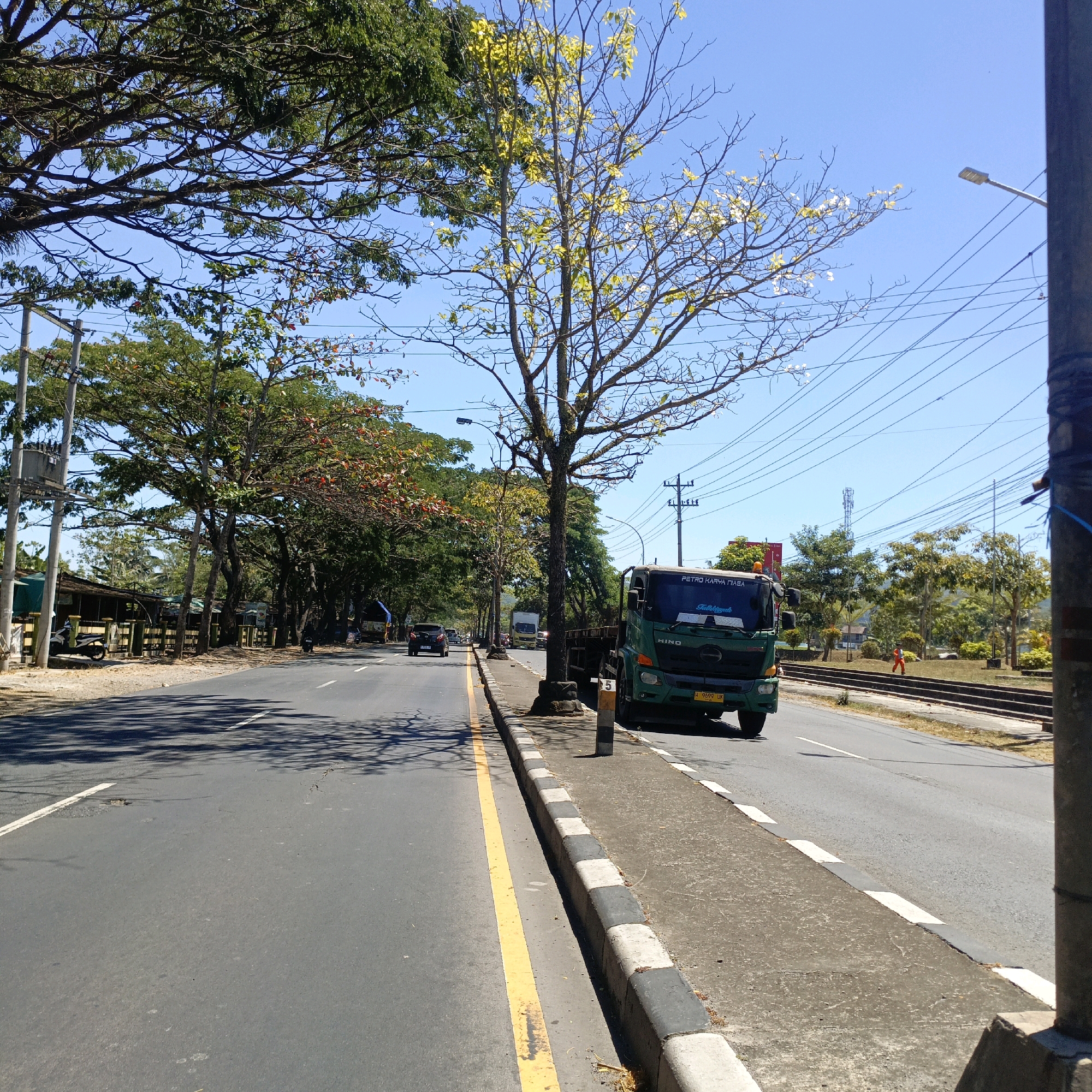The image depicts a road scene in Salatiga, Indonesia, on a clear, sunny day. A green Hino truck with the license plate H 9699 UK is prominent in the right foreground, driving on the road. The road is paved and has a yellow line marking the lane separation on the left and white dashed lines on the right. A concrete sidewalk with alternating black and white curb stones runs alongside the road. To the left of the road, there are several trees with green foliage, some appearing to be mature. A few vehicles, including a dark car, are visible further down the road. On the far left, there are some buildings with awnings and parked motorcycles. In the background, there is a telephone pole with numerous electrical wires, a street light, and more trees, with a cell tower visible in the distance. To the right of the road, there are railroad tracks and a grassy embankment with some trees. A person in an orange uniform is walking on the embankment. The overall environment suggests a tropical or subtropical climate with abundant vegetation.