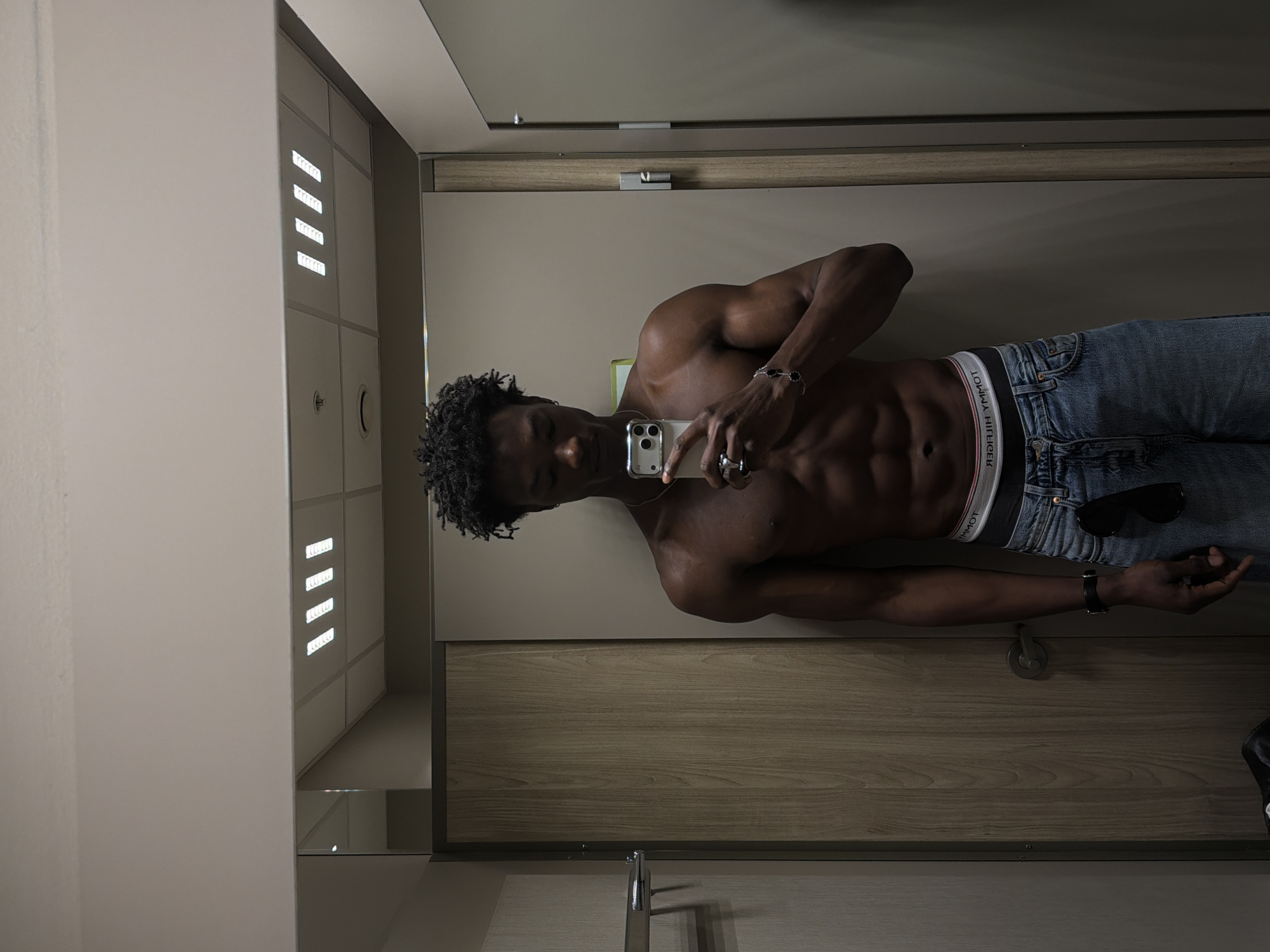 The image shows a young Black man with dark, curly hair, standing shirtless in what appears to be a fitting room or changing room. He is holding a smartphone in his right hand, taking a selfie in a mirror that reflects the ceiling lights and part of the room. He is wearing blue jeans and white and black underwear. Visible in the reflection of the mirror are several linear fluorescent light fixtures on a white suspended ceiling. The room has wooden paneling on the left wall and a plain gray wall on the right. A metal bar is mounted on the wooden wall. The man's physique is muscular, with visible abdominal muscles. He is wearing rings on his fingers and a bracelet on his left wrist. The image does not contain any information to suggest the location is in London, United Kingdom.