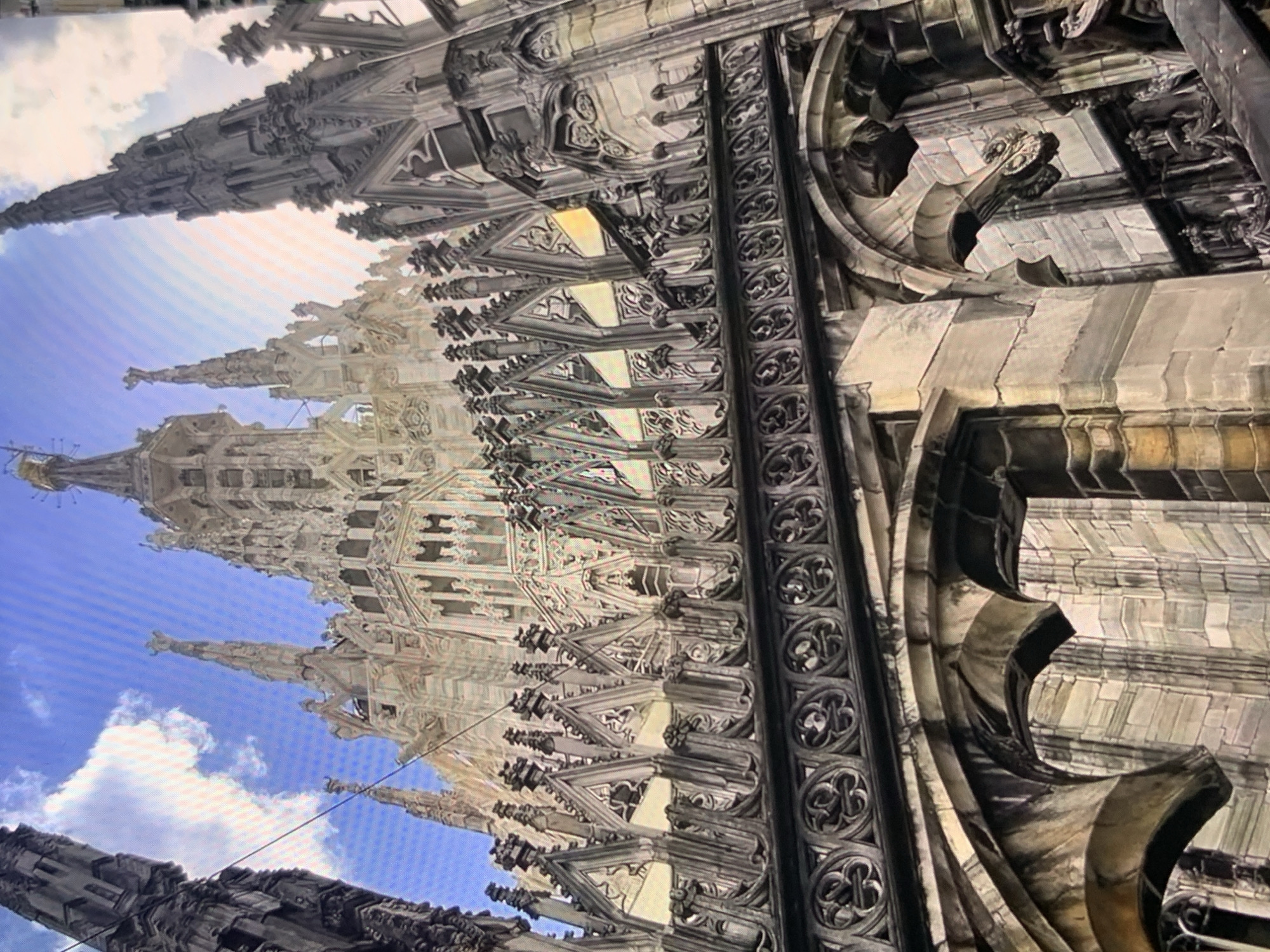 The image displays a close-up view of the intricate spires and Gothic architecture of a large cathedral, likely the Duomo di Milano, given its distinctive style. The perspective is from a low angle, looking upwards towards the sky. Ornate stone carvings, pointed arches, and decorative railings are visible. The sky is blue with scattered white clouds. The image does not depict the city of Stavropol, Russia, nor does it contain any people or specific actions.