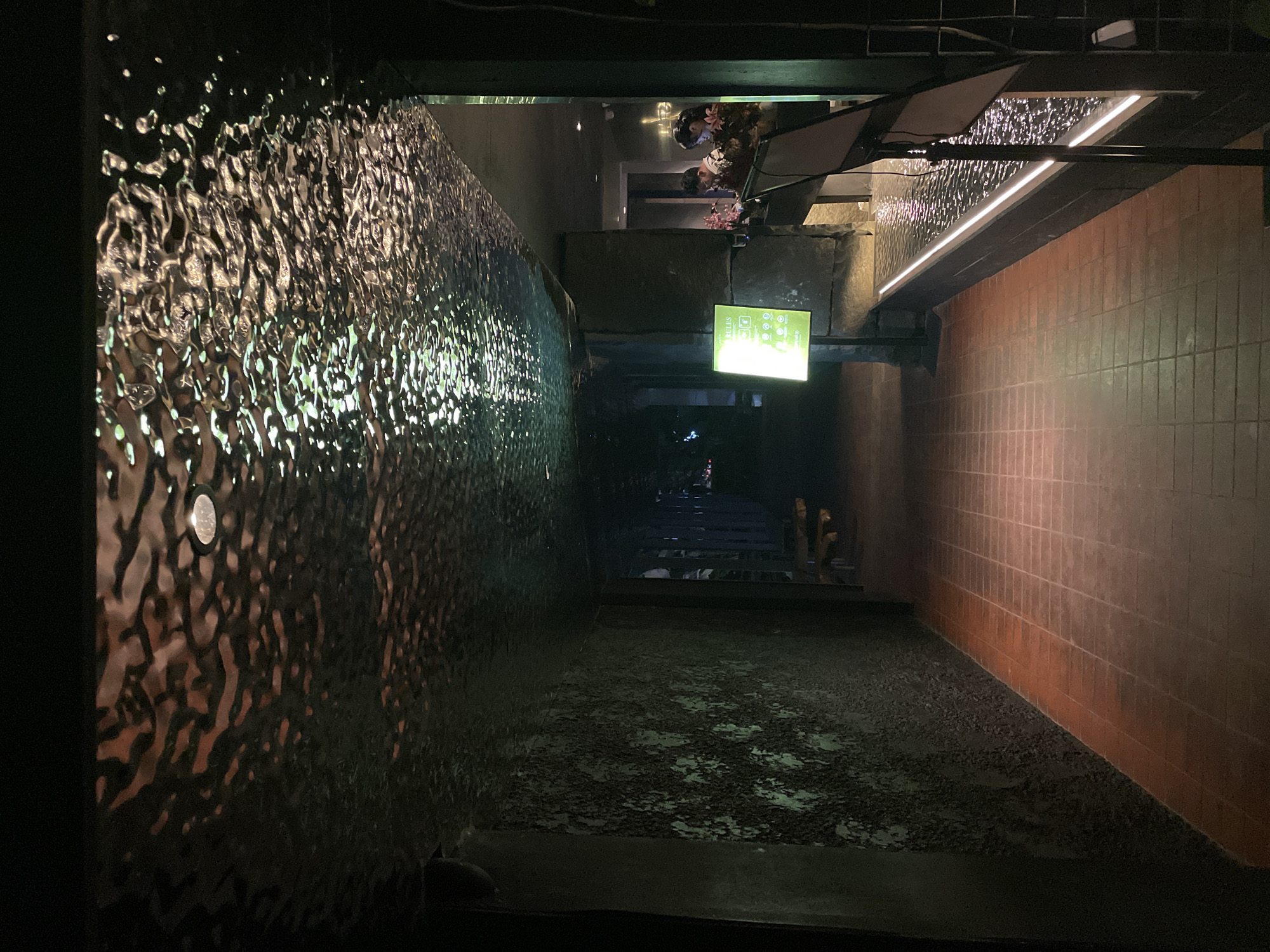 The image depicts an indoor walkway in South Jakarta, Indonesia, possibly a restaurant or upscale establishment. The left wall features a textured, possibly water-covered surface, reflecting light. On the right, there is a tiled wall in shades of orange. The floor of the walkway appears to be dark with a rough texture. Overhead, there are pipes, lights, and what appears to be a menu board. In the distance, there are blurry figures of people. The perspective is from a low angle, looking into the dimly lit walkway.