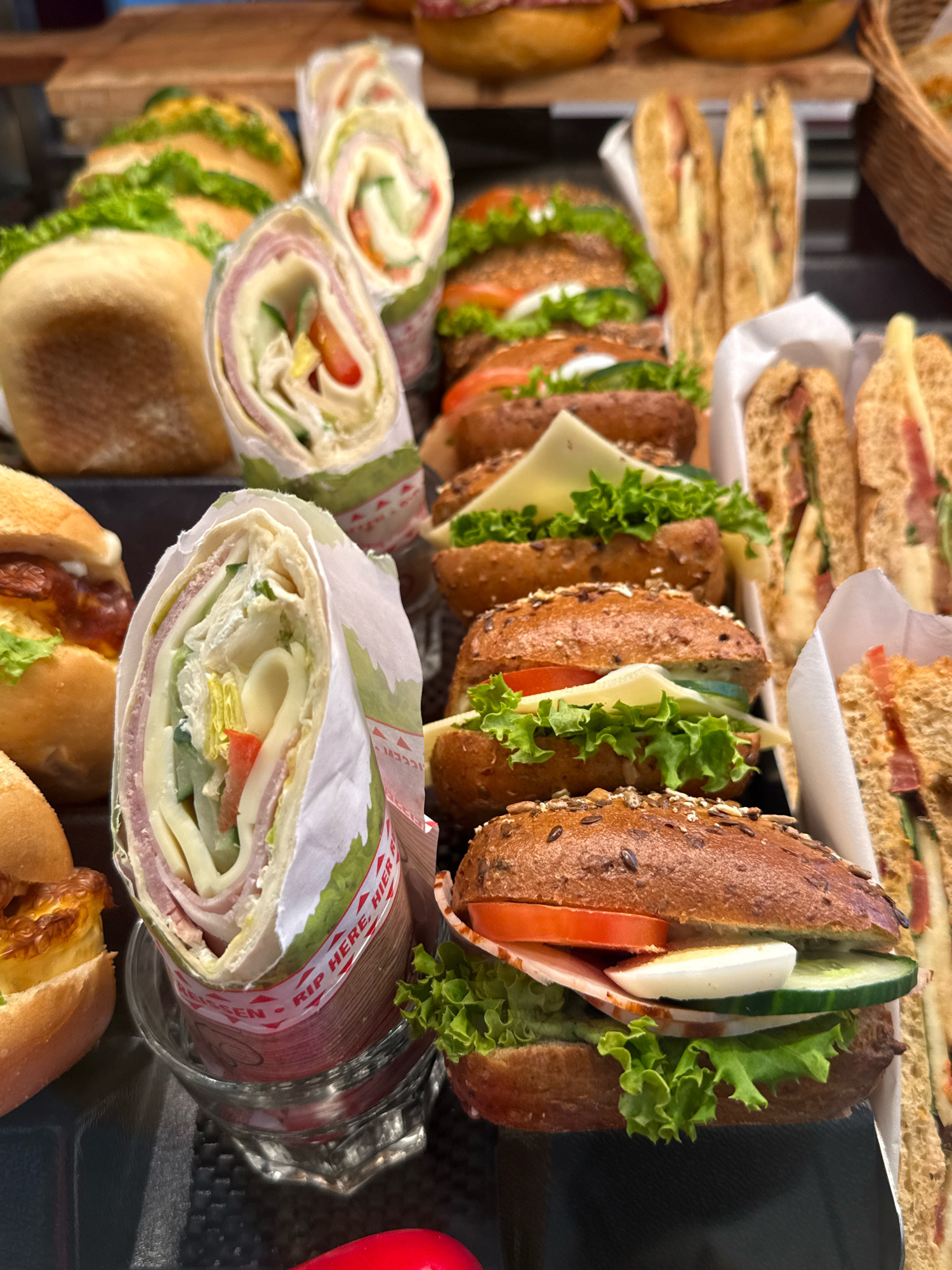 The image shows a close-up, overhead view of a variety of sandwiches and wraps displayed on a dark surface. In the foreground, a whole wheat bun sandwich is visible, layered with lettuce, tomato, ham, a slice of hard-boiled egg, and cucumber. To its left, a partially unwrapped wrap, filled with lettuce, cheese, ham, and tomato, sits in a clear glass container. Behind these items, several other sandwiches and wraps are arranged. Some appear to be halved sandwiches on whole wheat bread, while others are rolled wraps. In the background, a basket is partially visible on the right. The lighting is bright, illuminating the food items. The setting appears to be a food display or counter.